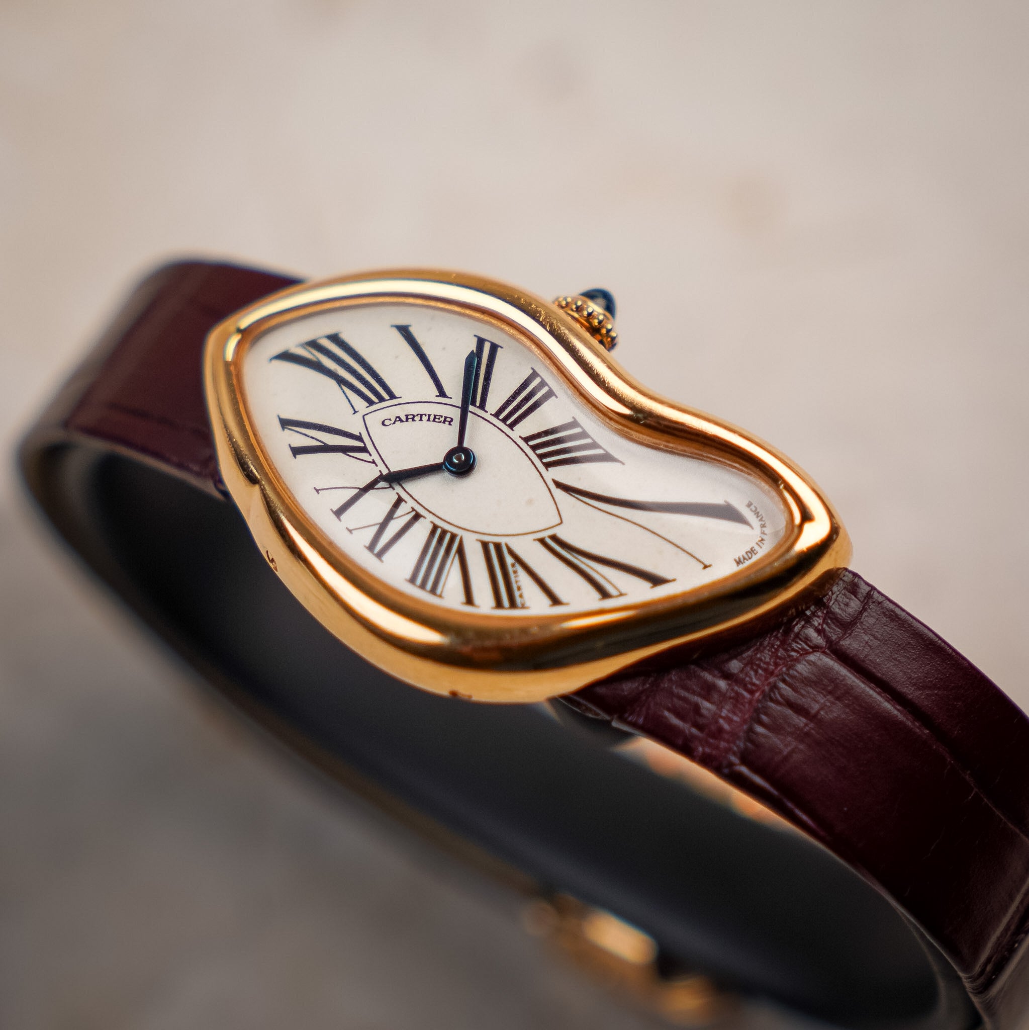 A close-up of a Cartier watch with a gold-tone heart-shaped bezel, white dial with black Roman numerals, and a dark burgundy leather strap on a neutral beige background.