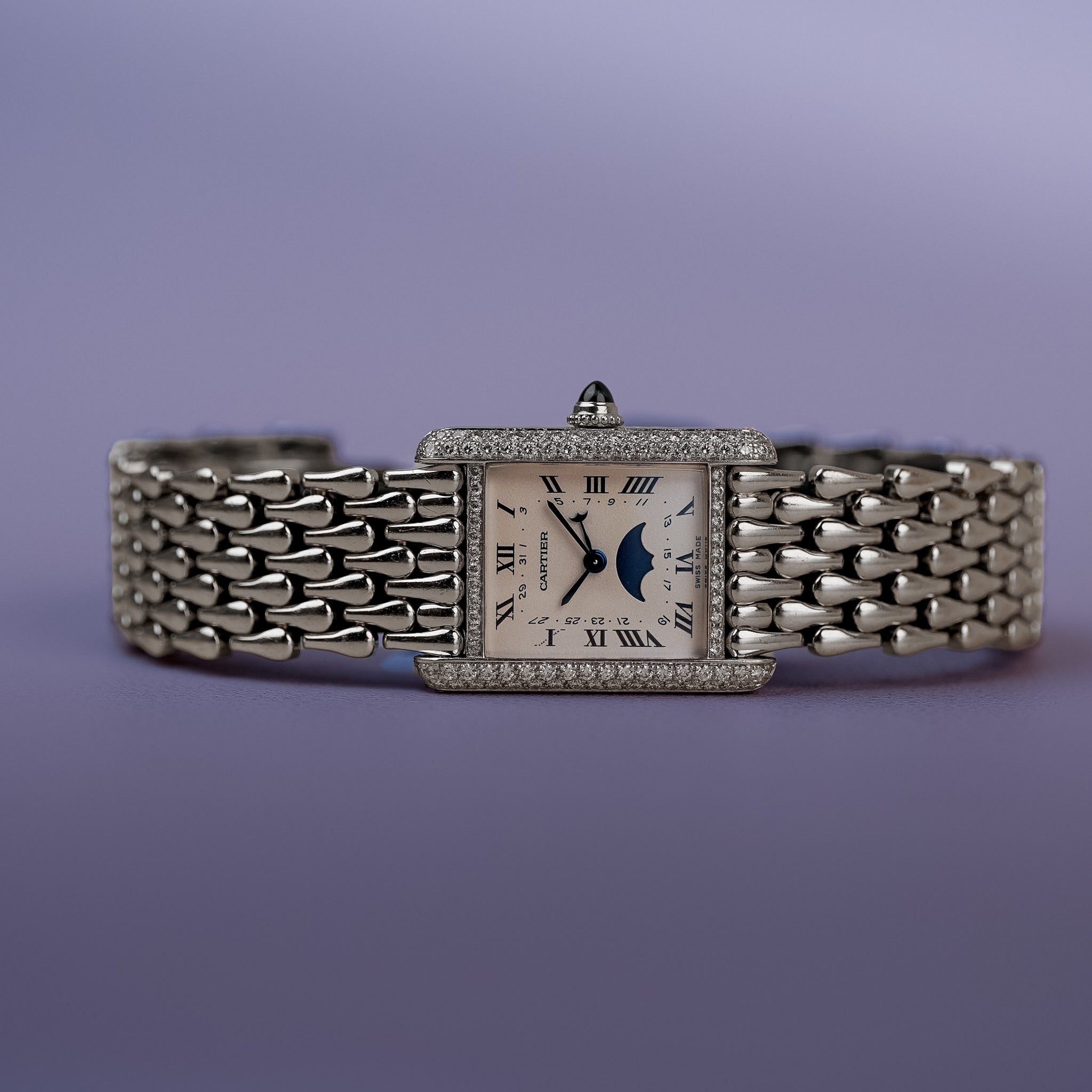 A white gold Cartier Tank with white dial, Roman numerals, a moonphase indicator, diamond bezel, and a white gold bracelet. Lying sideways on a purple surface