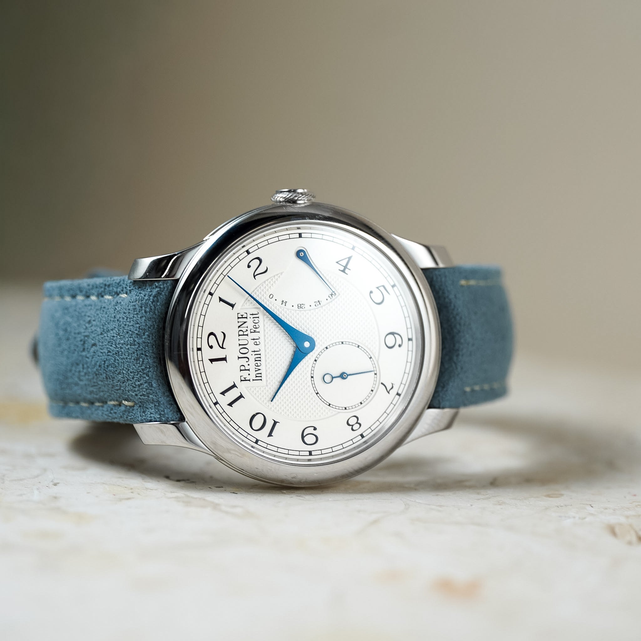 A close-up shot of a wristwatch with a silver-tone case, white dial, blue hands, and a blue suede strap, resting on a light textured surface.