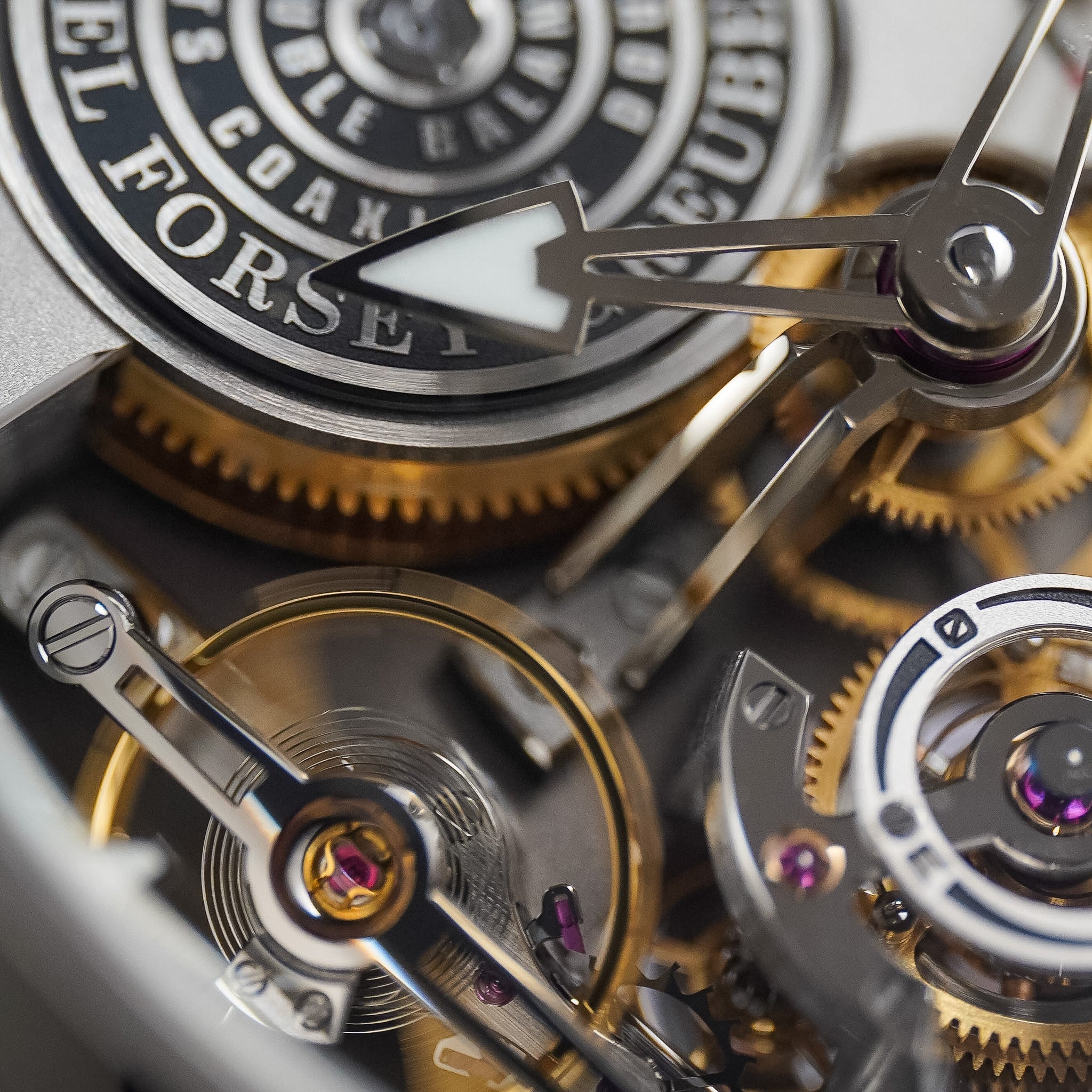 Extreme close-up of a mechanical watch movement showing silver and gold-toned gears, screws, and a black-and-silver dial with white text.
