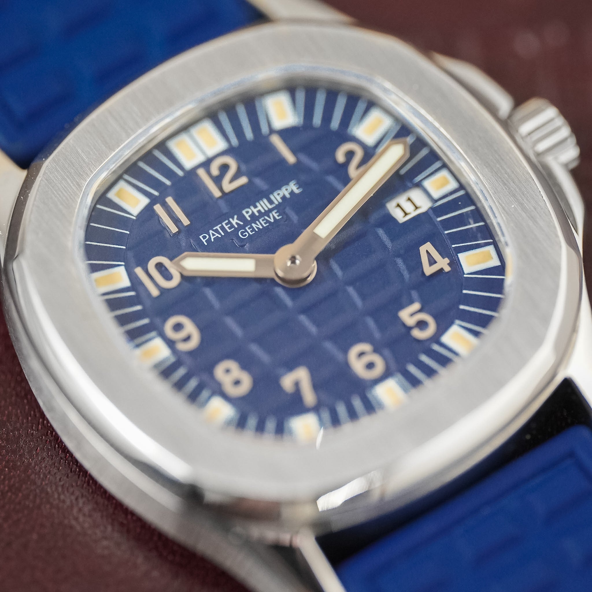 A closeup of a Patek Philippe ladies sports watch with a blue dial, blue strap, off-white hour markers and hands, and a steel case laying flat on a maroon surface.
