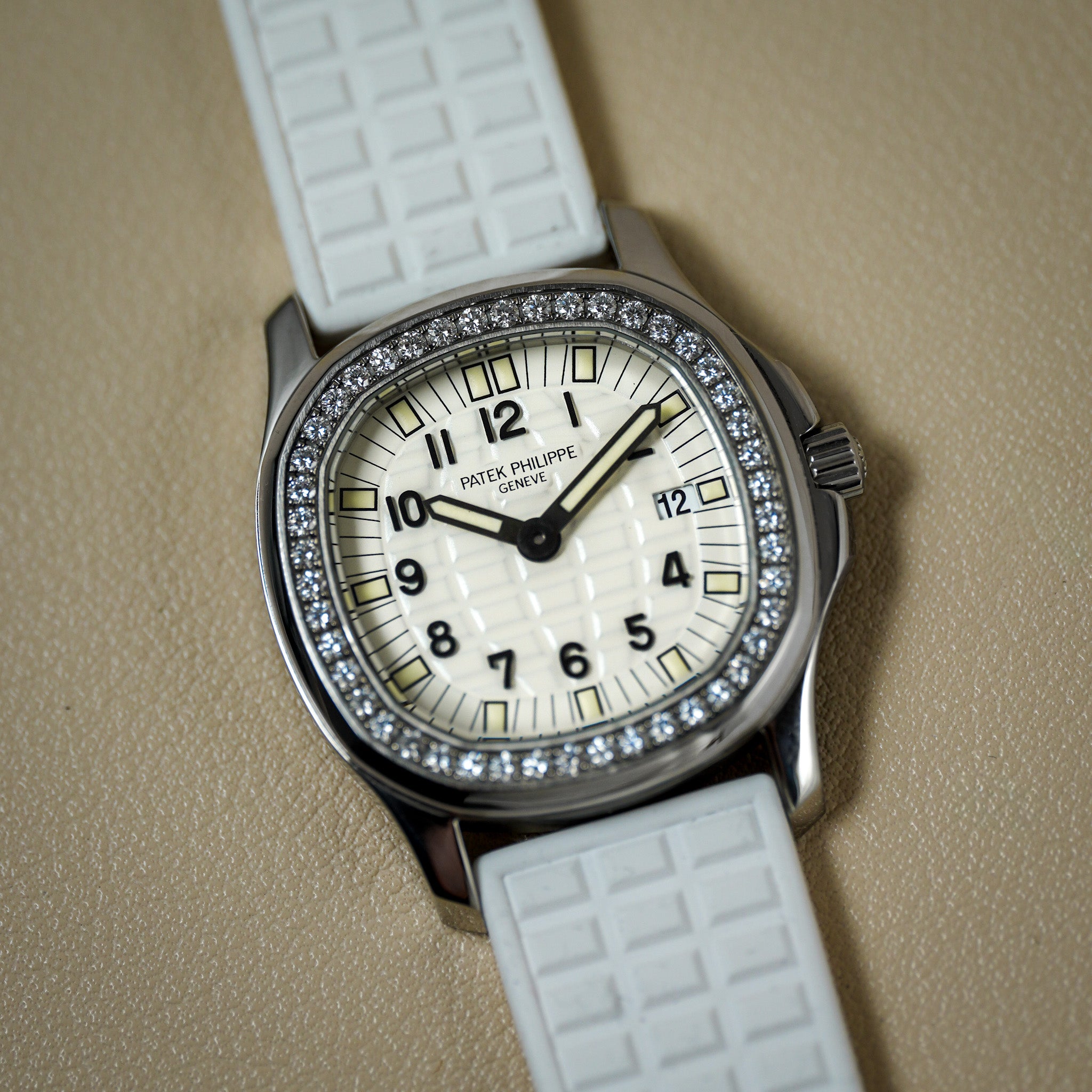 A white dial steel sport watch laying flat on a cream surface. The watch has a diamond bezel, white rubber strap, steel case, and black numerals for the hour markers.