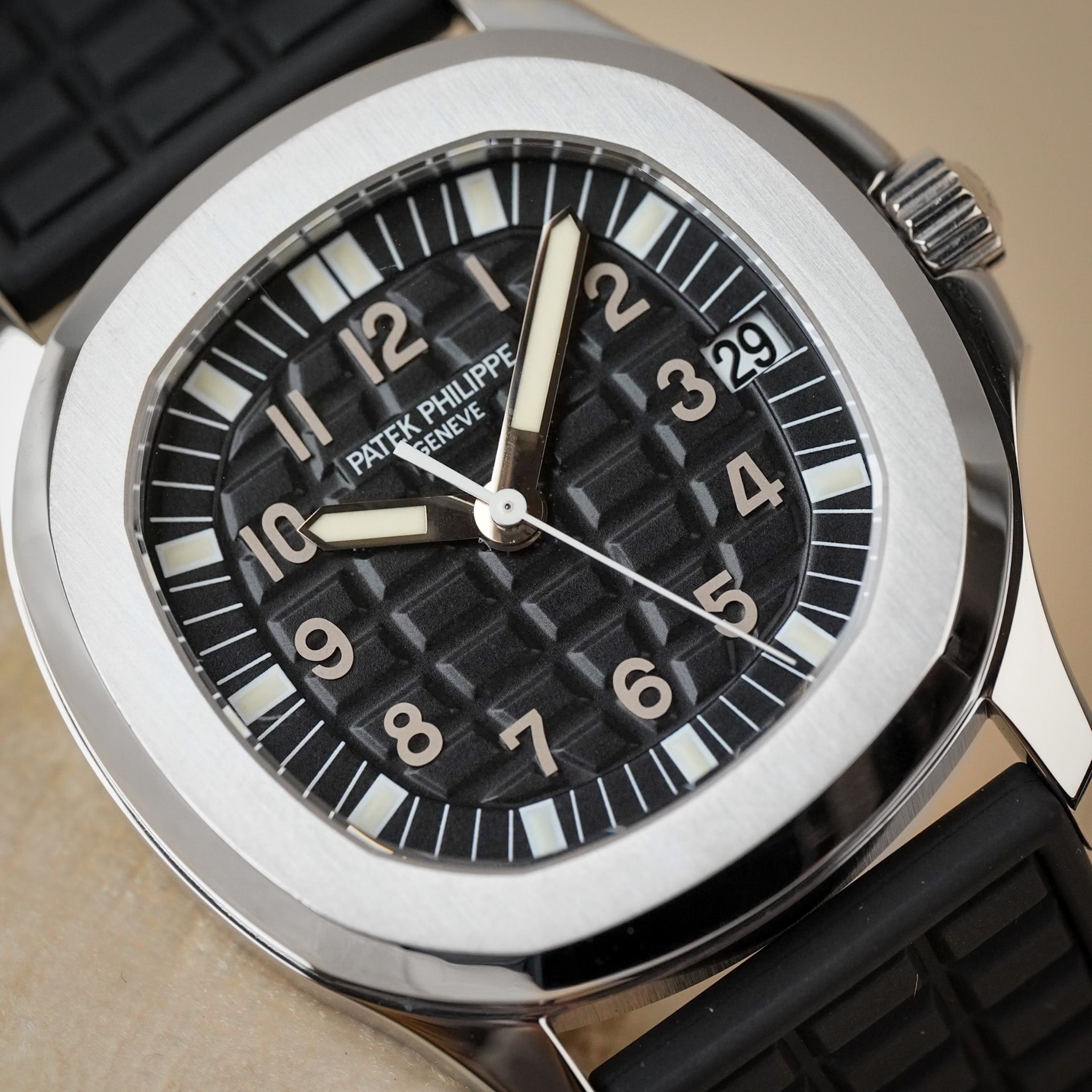 A stainless steel Patek Philippe Aquanaut sport watch with a textured black dial, textured black rubber strap, and white hands and hour markers, shot against a neutral cream background