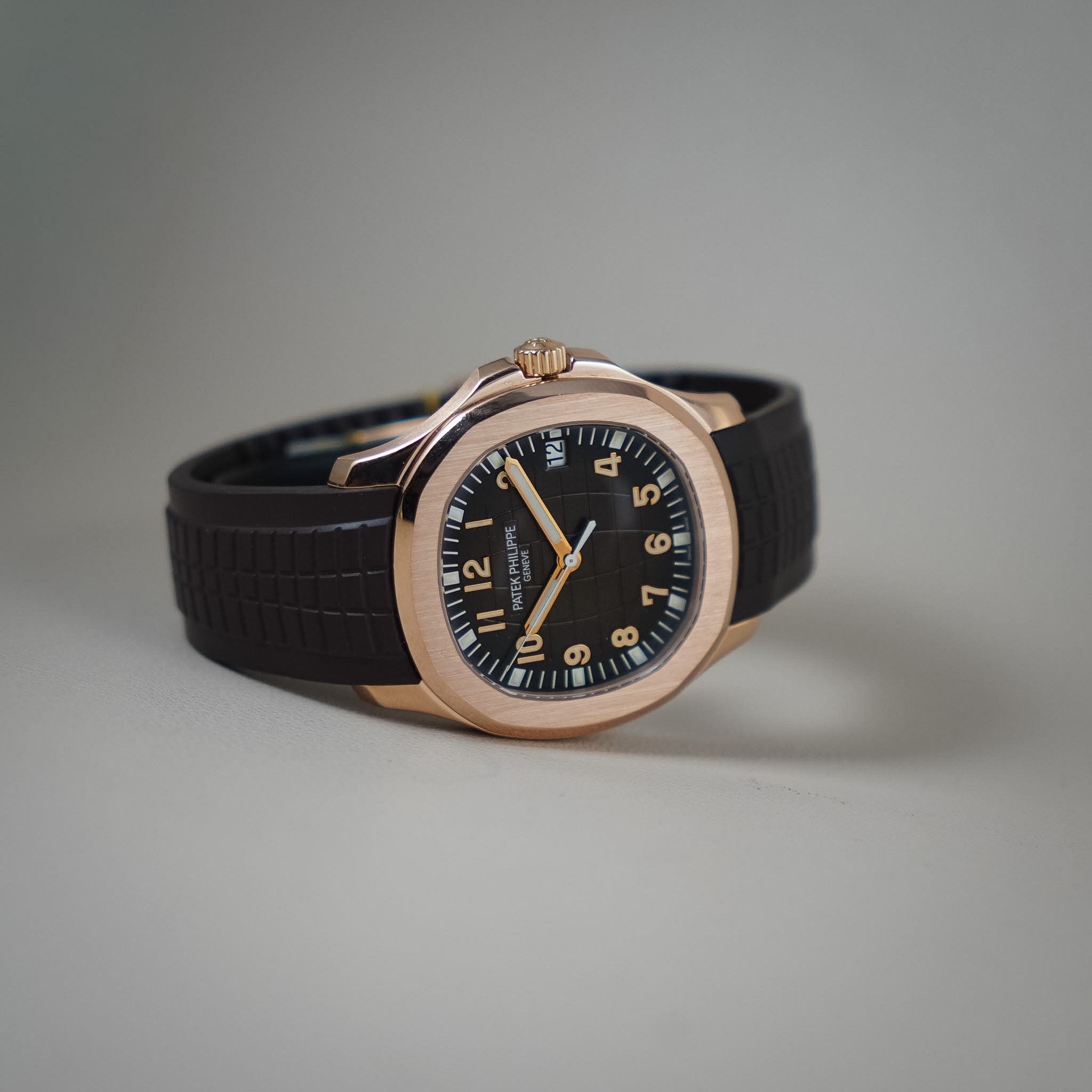 A Patek Philippe wristwatch with a rose gold-toned octagonal case, black dial, and black textured strap, photographed on a neutral gray background.