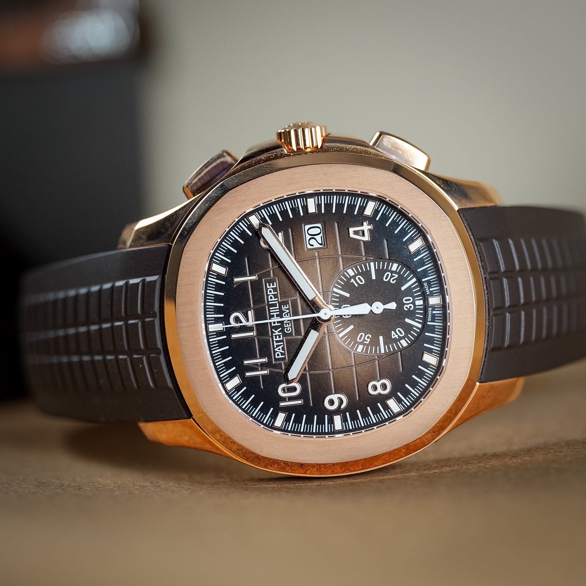 Photo of a luxury rose gold sport watch with a textured brown dial, brown rubber strap, and off-white hands and hour markers laying on its side on a tan table.
