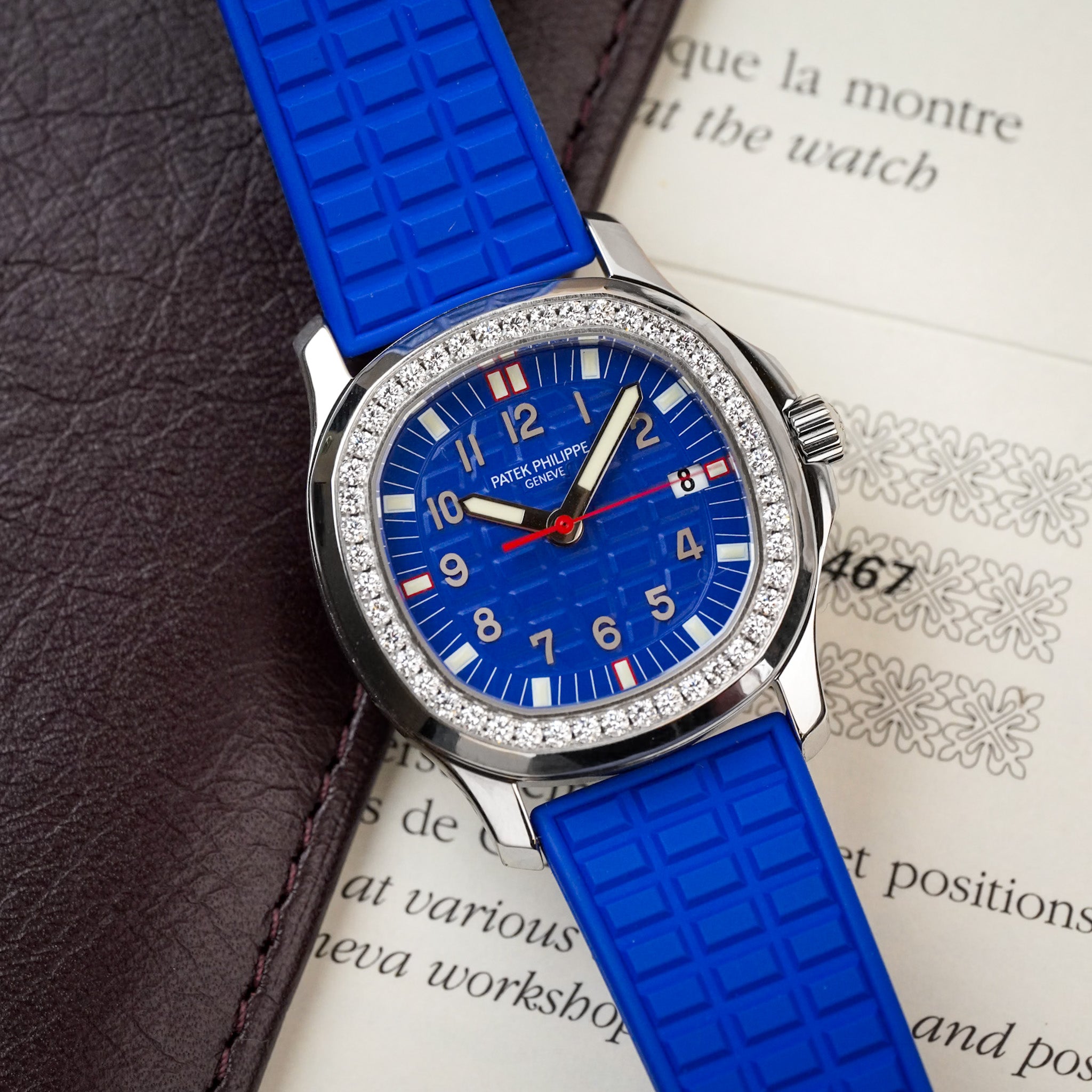 A stainless steel Patek Philippe Aquanaut with a textured blue dial featuring white arabic numerals and red accents, a textured blue rubber strap, and a diamond bezel, shot flat atop some papers and leather folio
