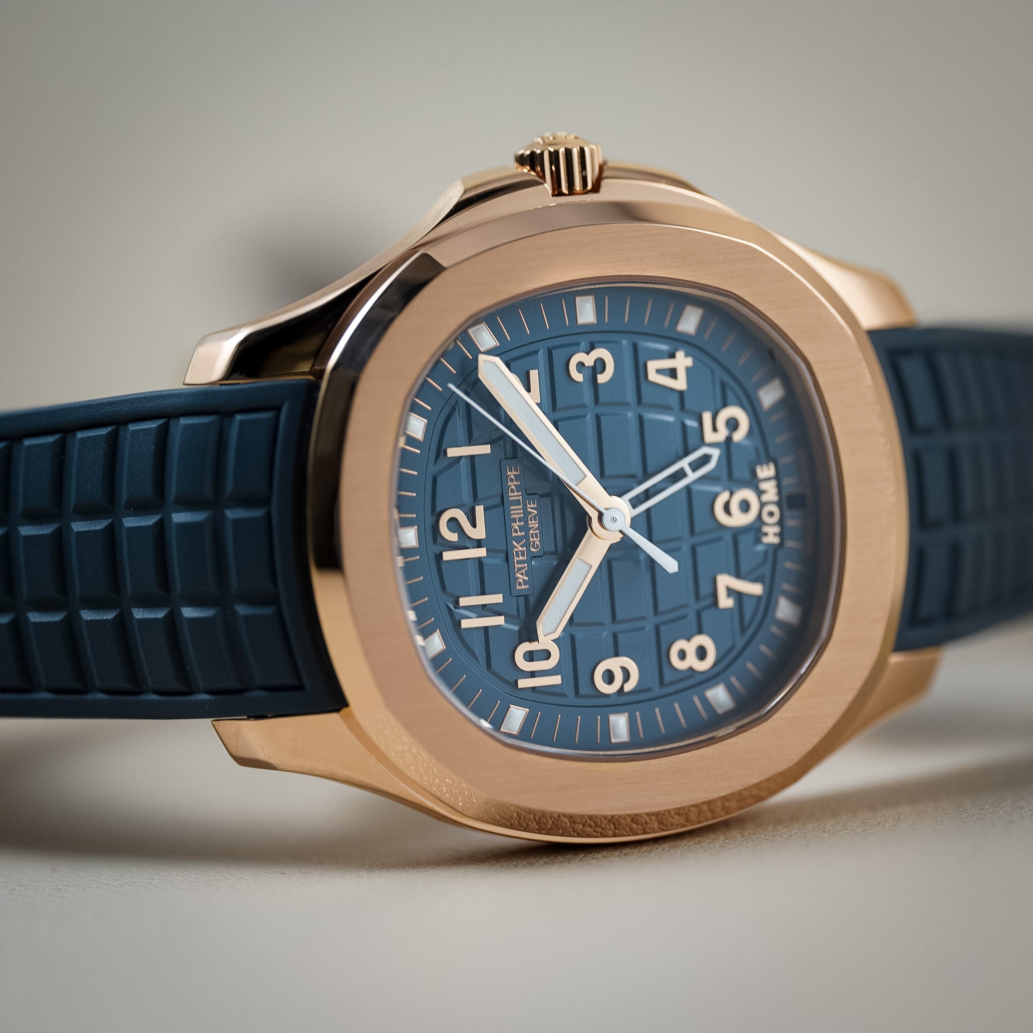 A close-up of a wristwatch with a blue dial, blue strap, and rose gold-toned case on a soft gray background.