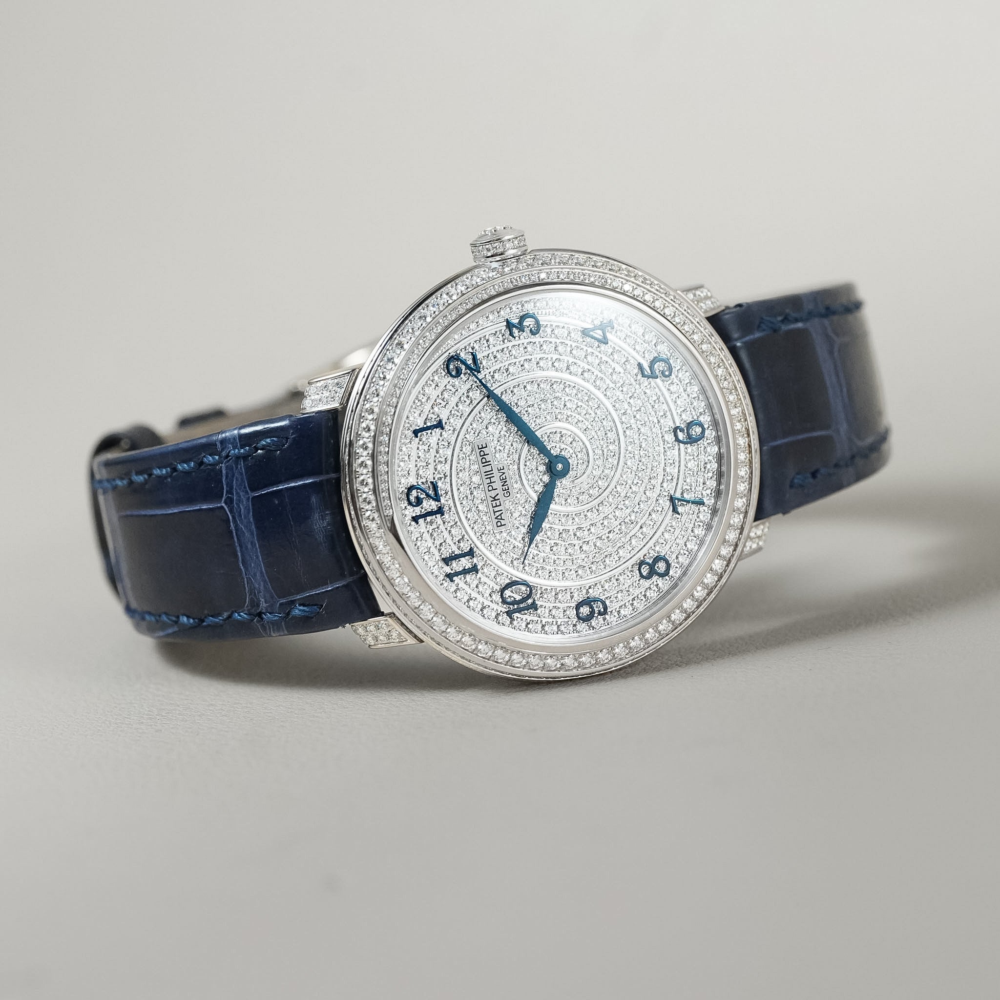 A luxury wristwatch with a silver-toned diamond-encrusted bezel, a diamond-set dial with blue Arabic numerals and hands, and a deep blue alligator leather strap, shown on a neutral light gray background.