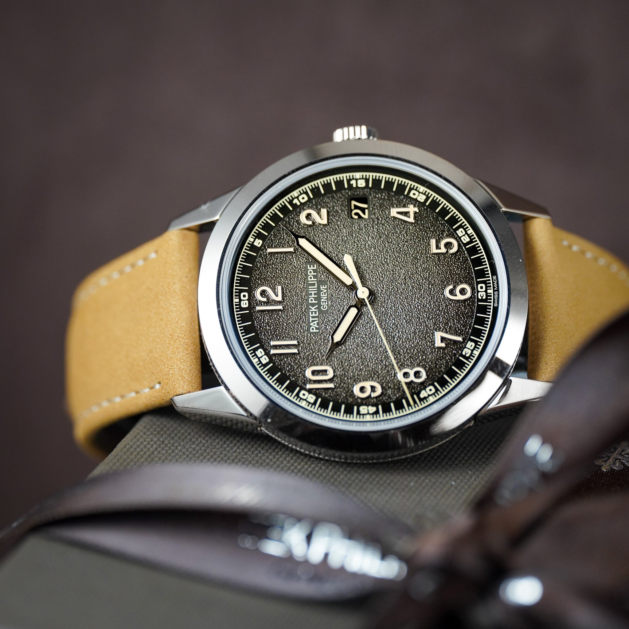 A sideways photo of a silver-toned watch with a tan strap, dark dial, and cream colored hands and hour marker against a dark, blurry background.