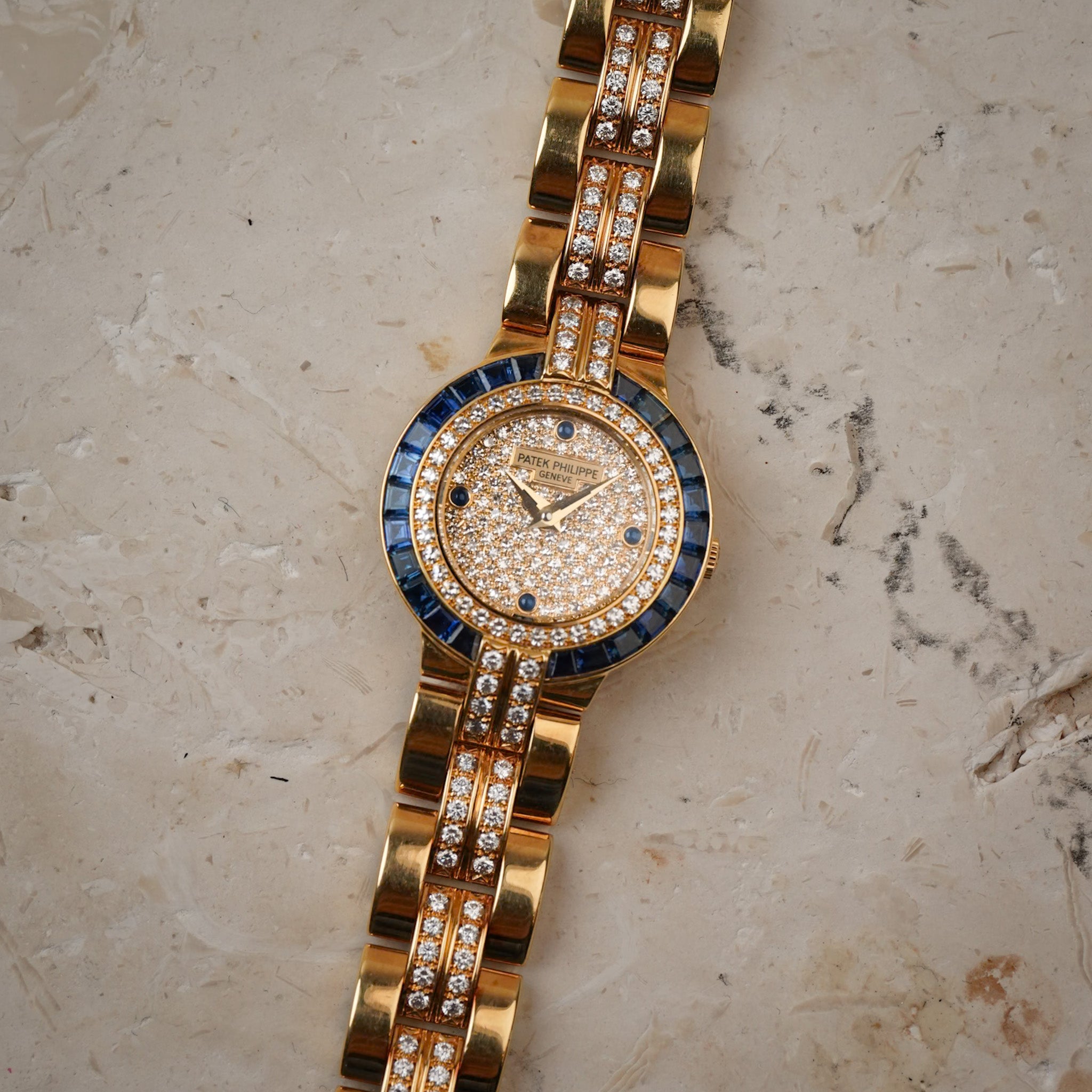 A yellow gold ladies wristwatch with a pavé diamond dial, blue sapphire bezel, a yellow gold and diamond bracelet, laying flat on a stone surface