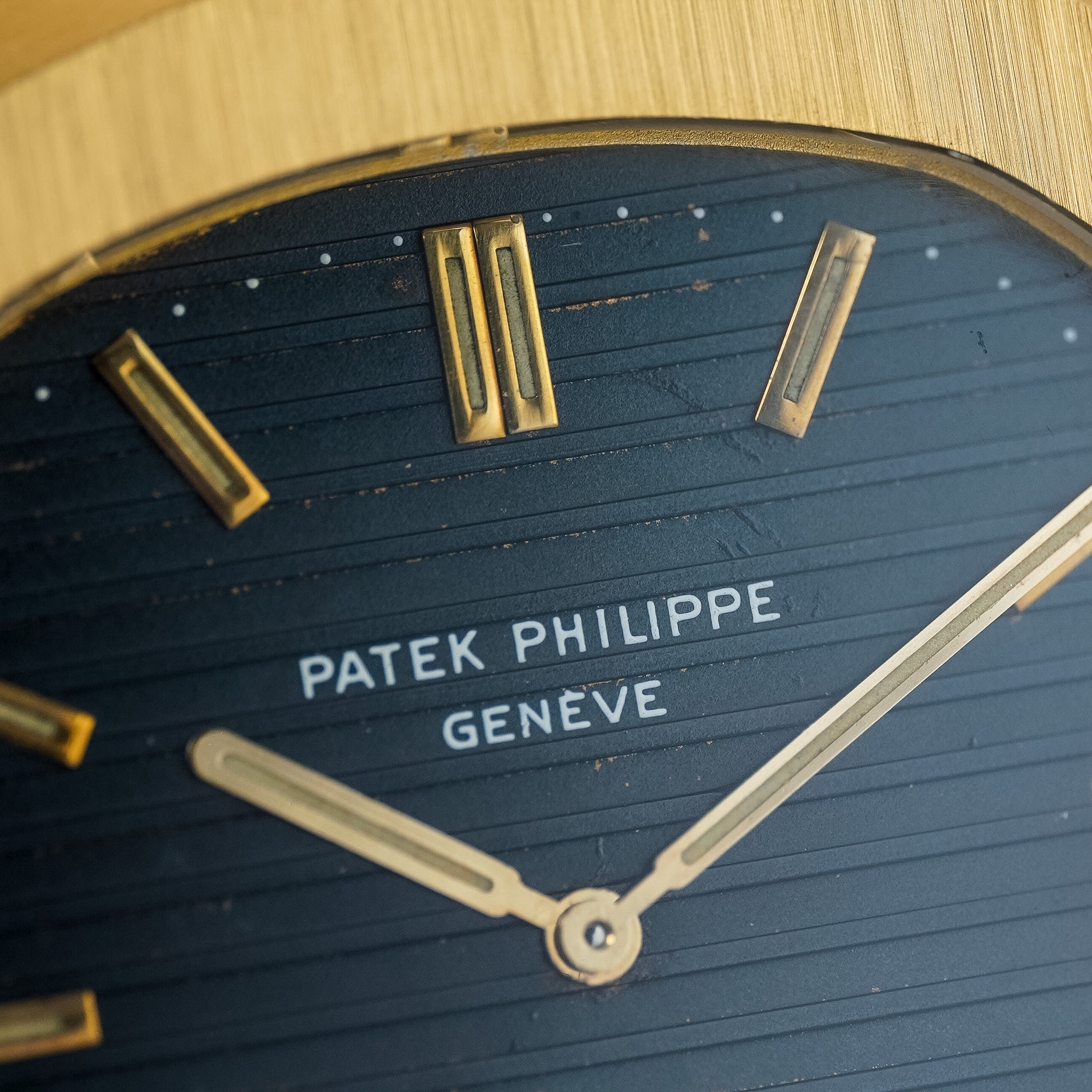 Close-up of a Patek Philippe watch dial with a dark blue textured background, gold-tone hour markers and hands, and the white Patek Philippe Geneva text at center.