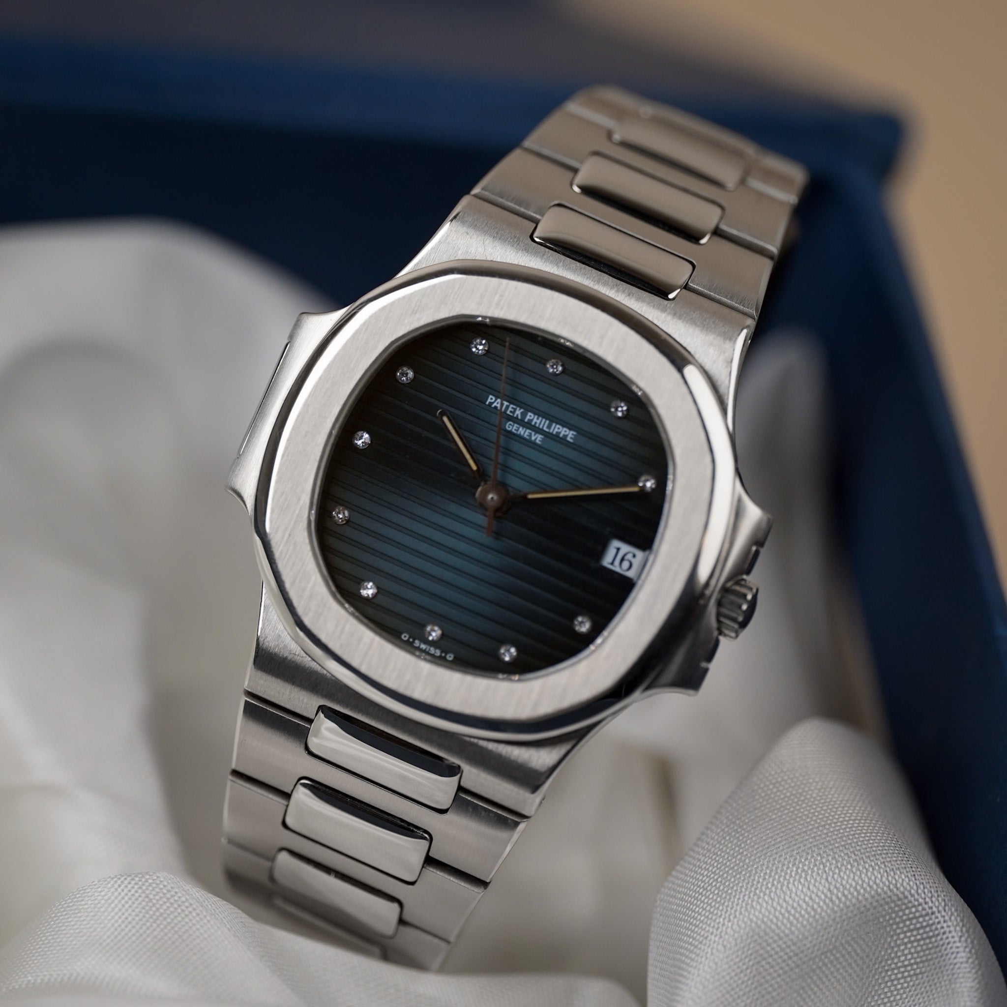 A steel Patek Philippe Nautilus with a ribbed blue dial, diamond hour markers, and a date window showing "16", shot against a white fabric and blue box backdrop