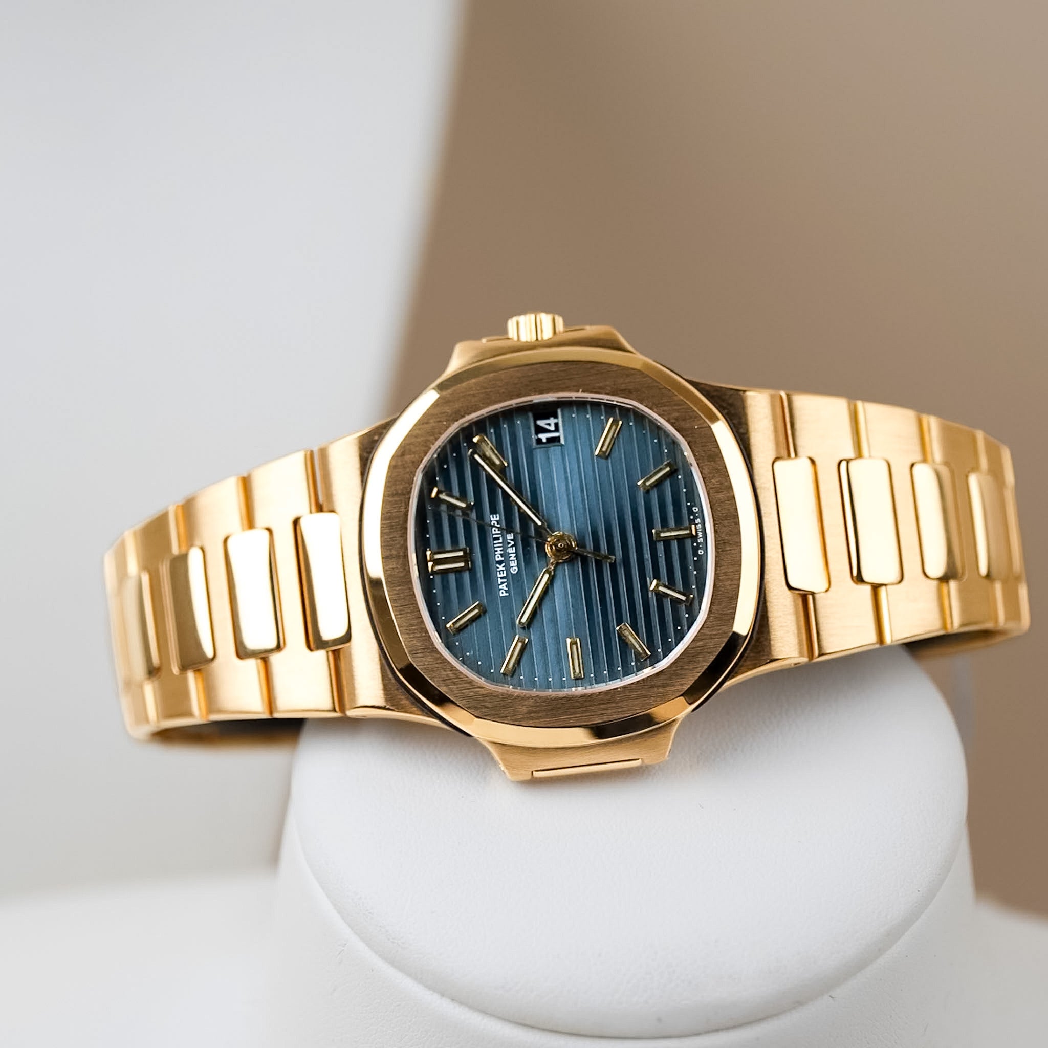 A yellow gold Patek Philippe Nautilus with a textured blue dial, gold hands and hour markers, a gold bracelet, and a date window showing "14", shot lying sideways on a white cylinder