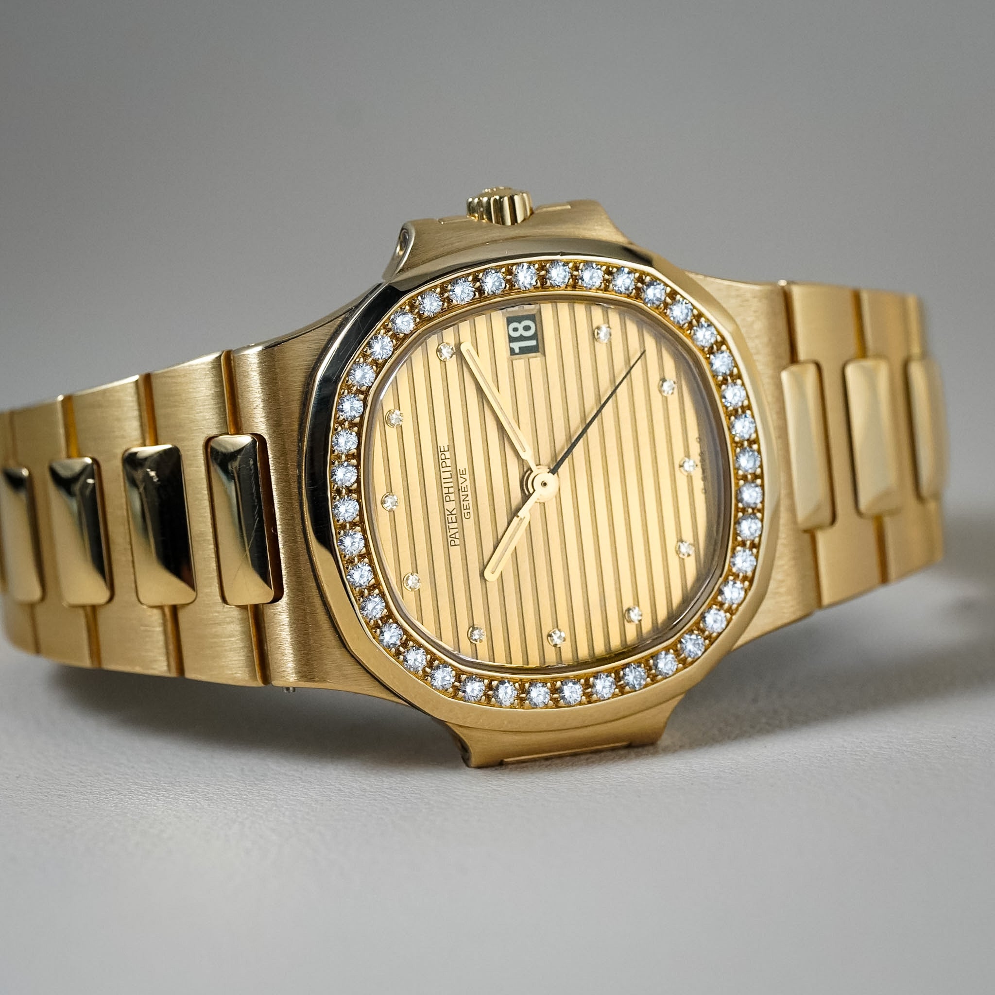 A gold wristwatch with a diamond-accented bezel, a gold striped dial, and a matching gold-tone metal link bracelet, photographed on a neutral gray background.