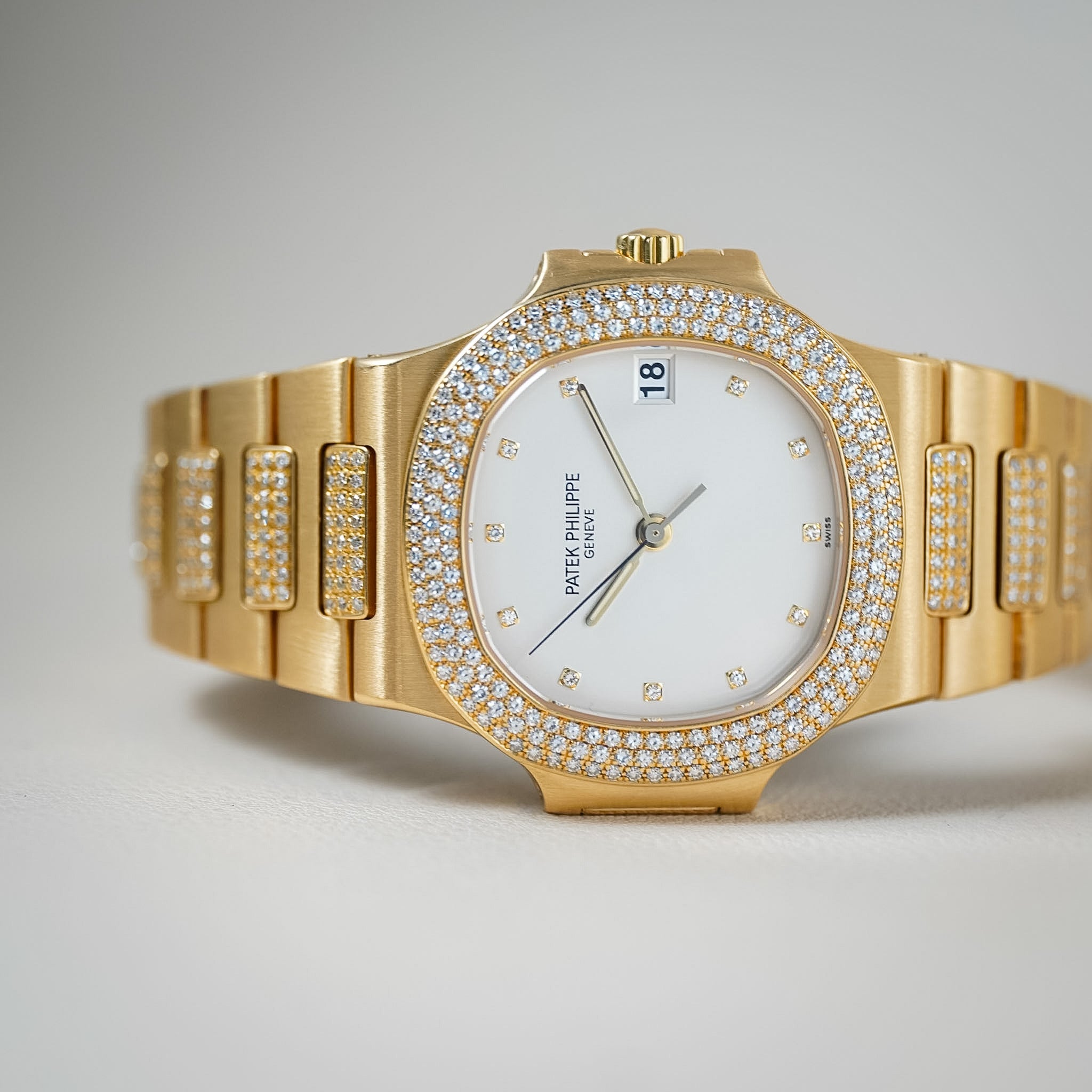 A gold-tone Patek Philippe Nautilus watch with a white dial, diamond-set bezel, and diamond-accented bracelet, photographed on a neutral light gray background.