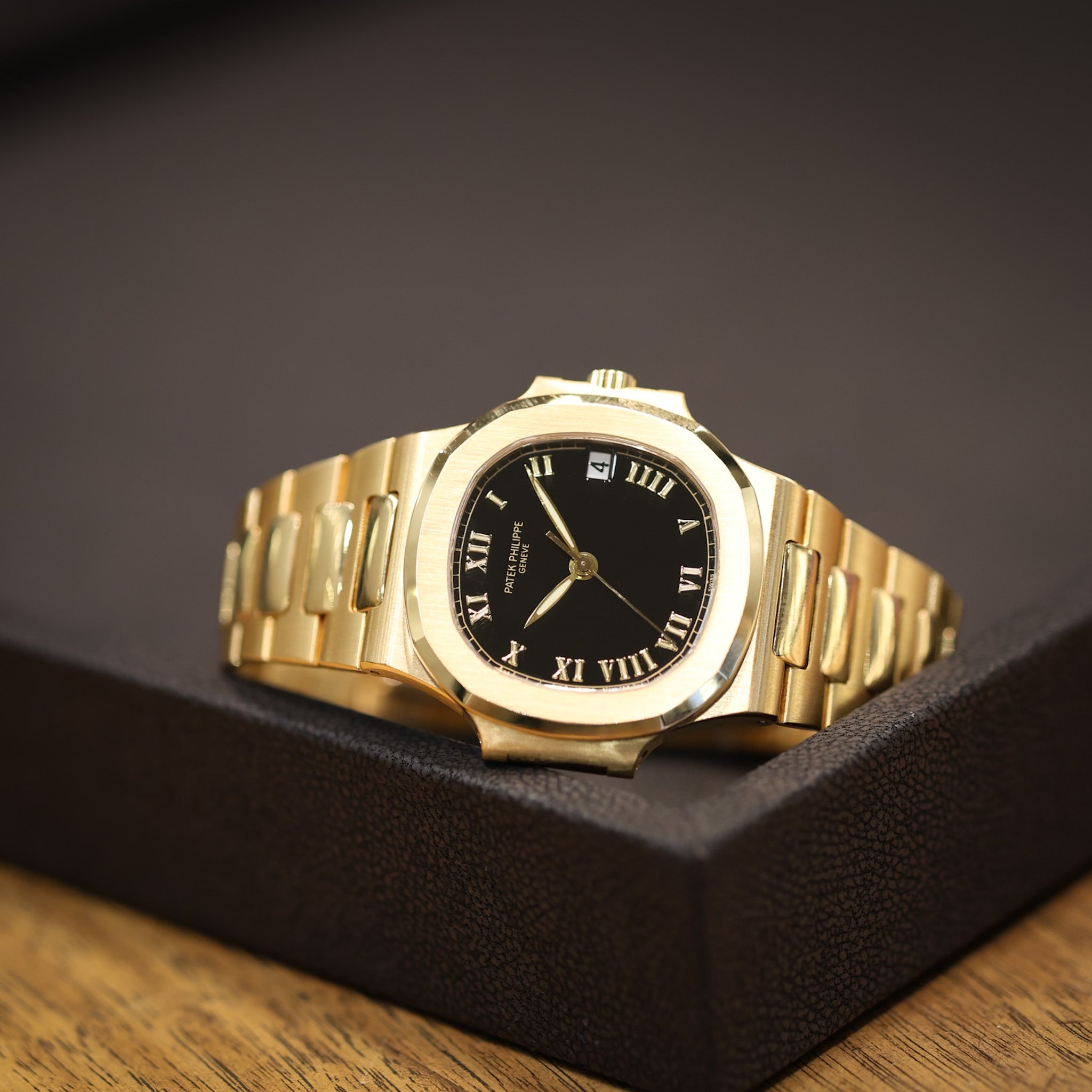 A gold-tone wristwatch with a black dial and Roman numeral hour markers, resting on a textured black display box over a wooden surface.