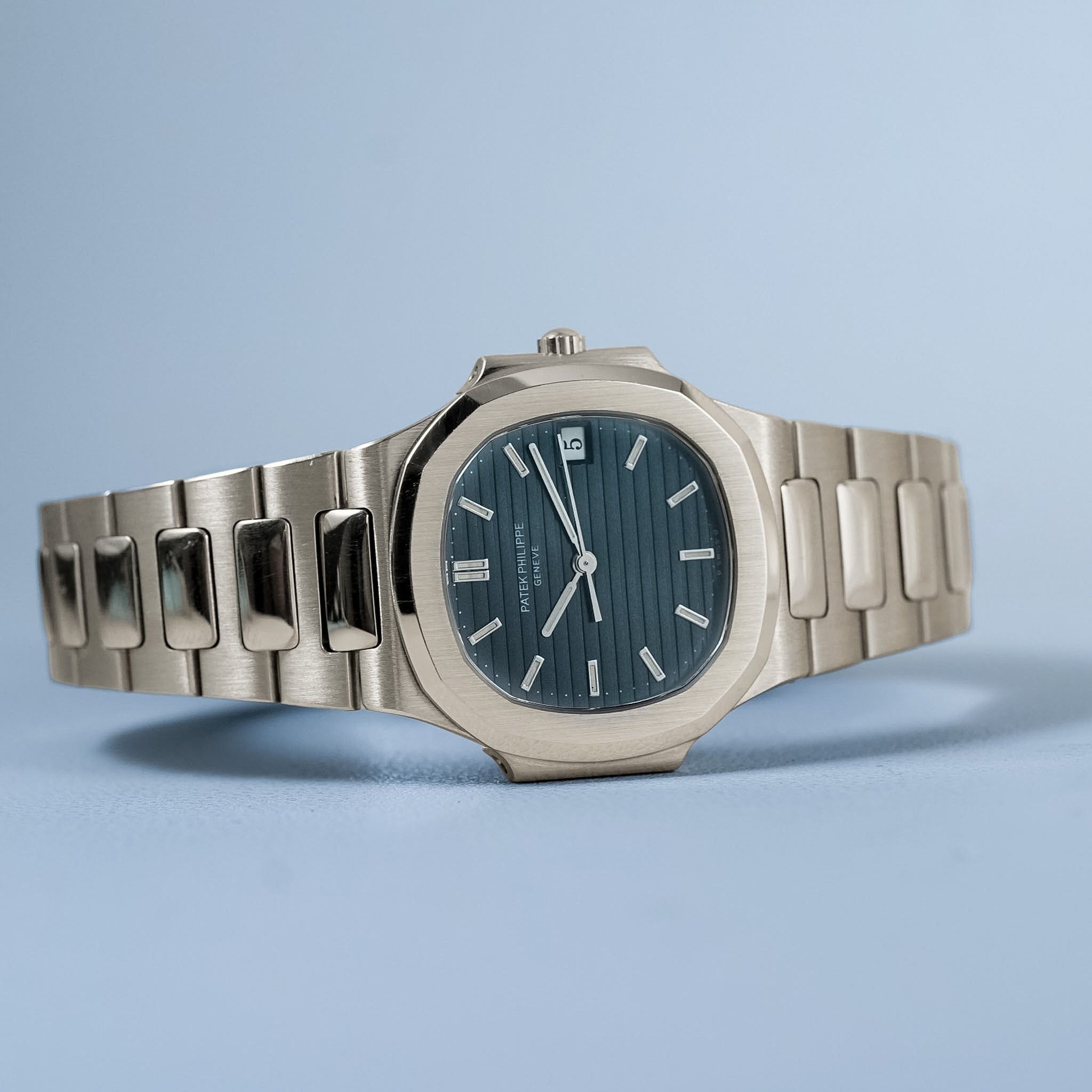A white gold watch laying sideways with a blue dial and integrated bracelet against a light blue backdrop.
