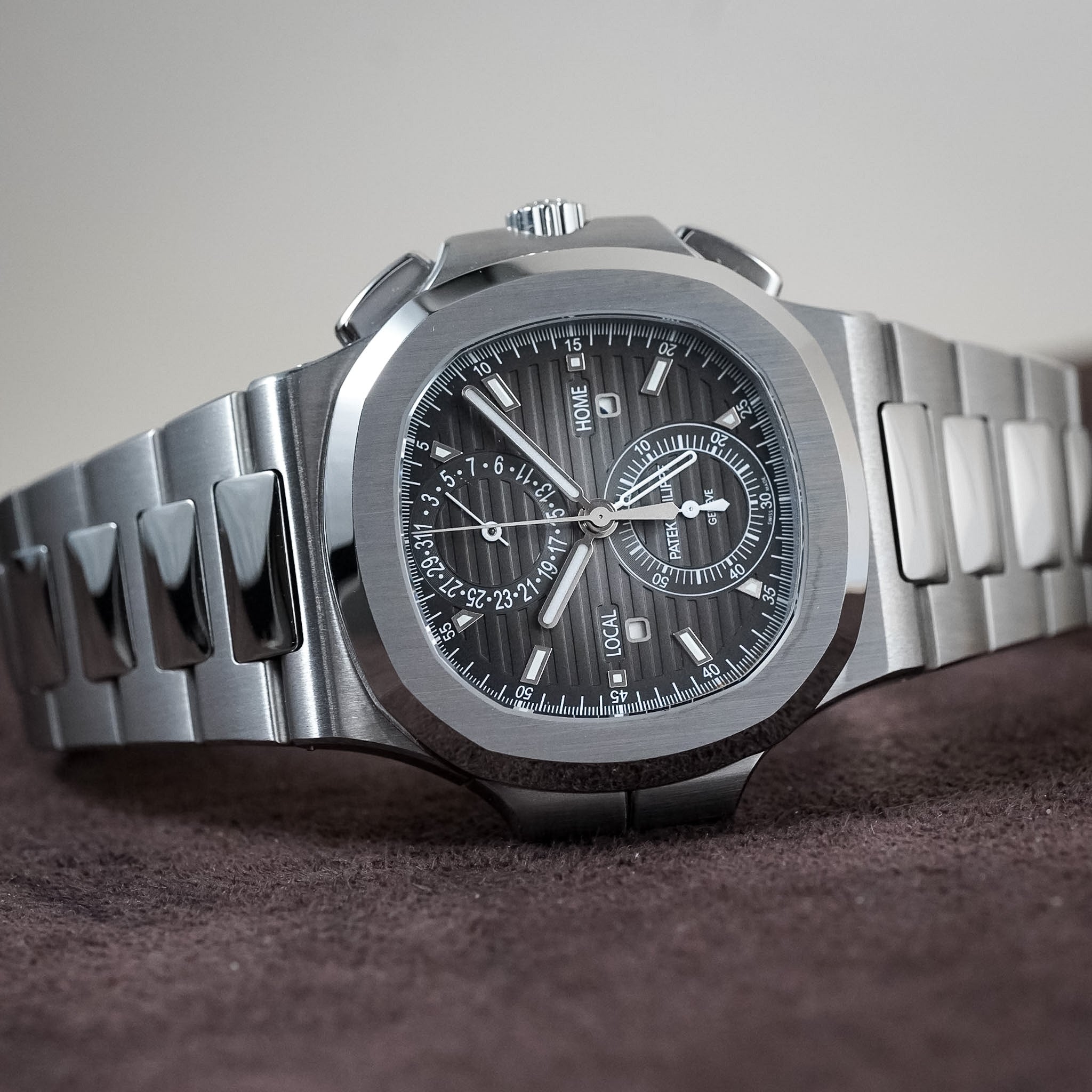 A stainless steel chronograph watch with a silver-toned metal link bracelet, featuring a dark gray sunburst dial with white hands and markers, resting on a dark brown textured surface against a neutral gray background.