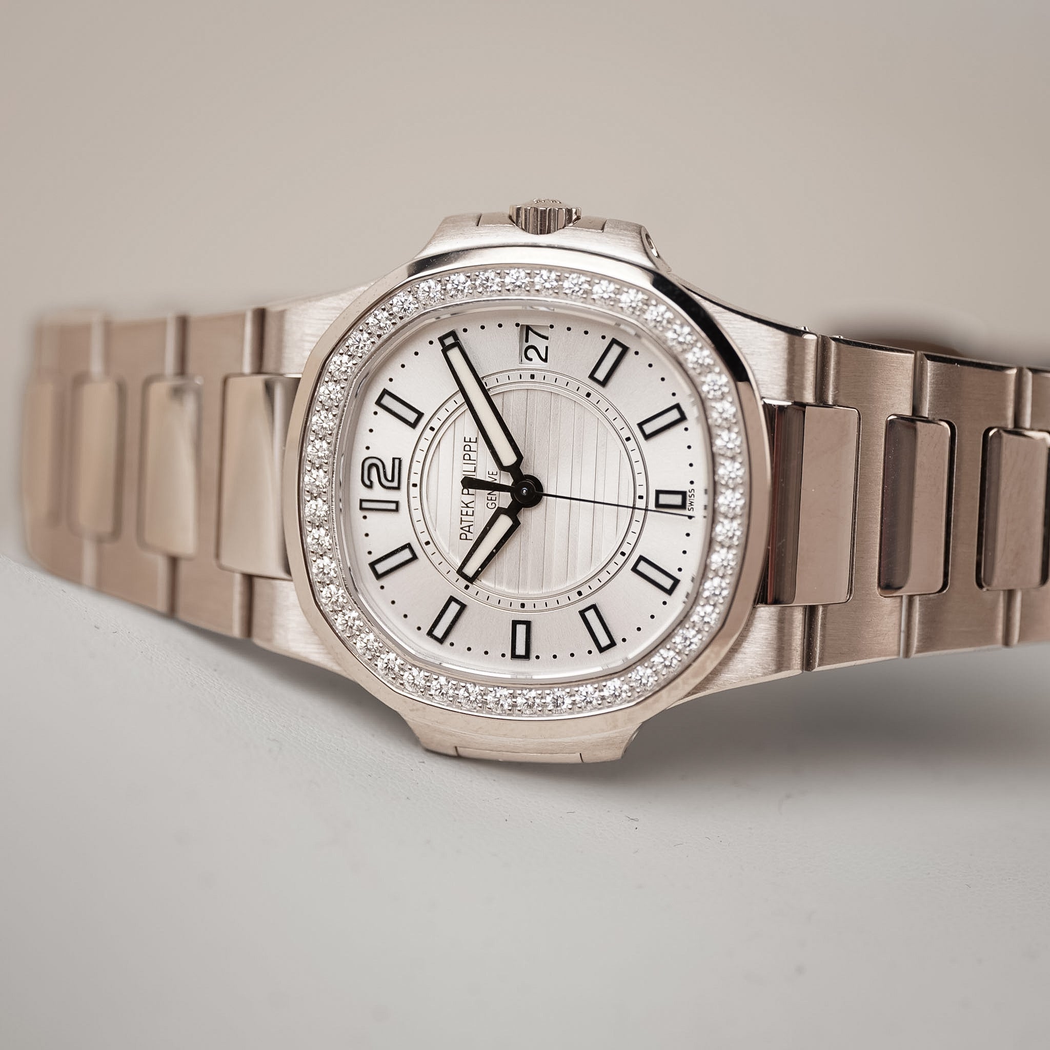A white gold ladies Patek Philippe Nautilus with a white dial, diamond bezel, and white gold bracelet. Shot resting sideways on a light grey surface.