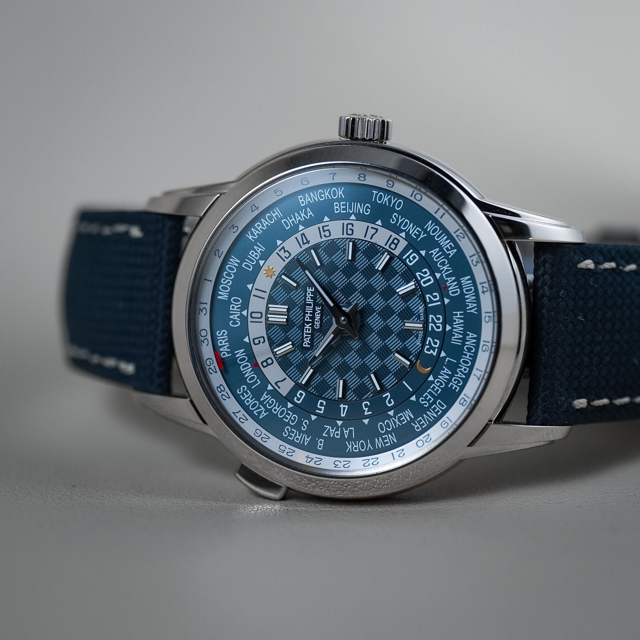 A close-up shot of a wristwatch with a blue and silver checkered dial, a silver stainless steel case, and a dark blue canvas strap with white stitching, set against a neutral gray background.