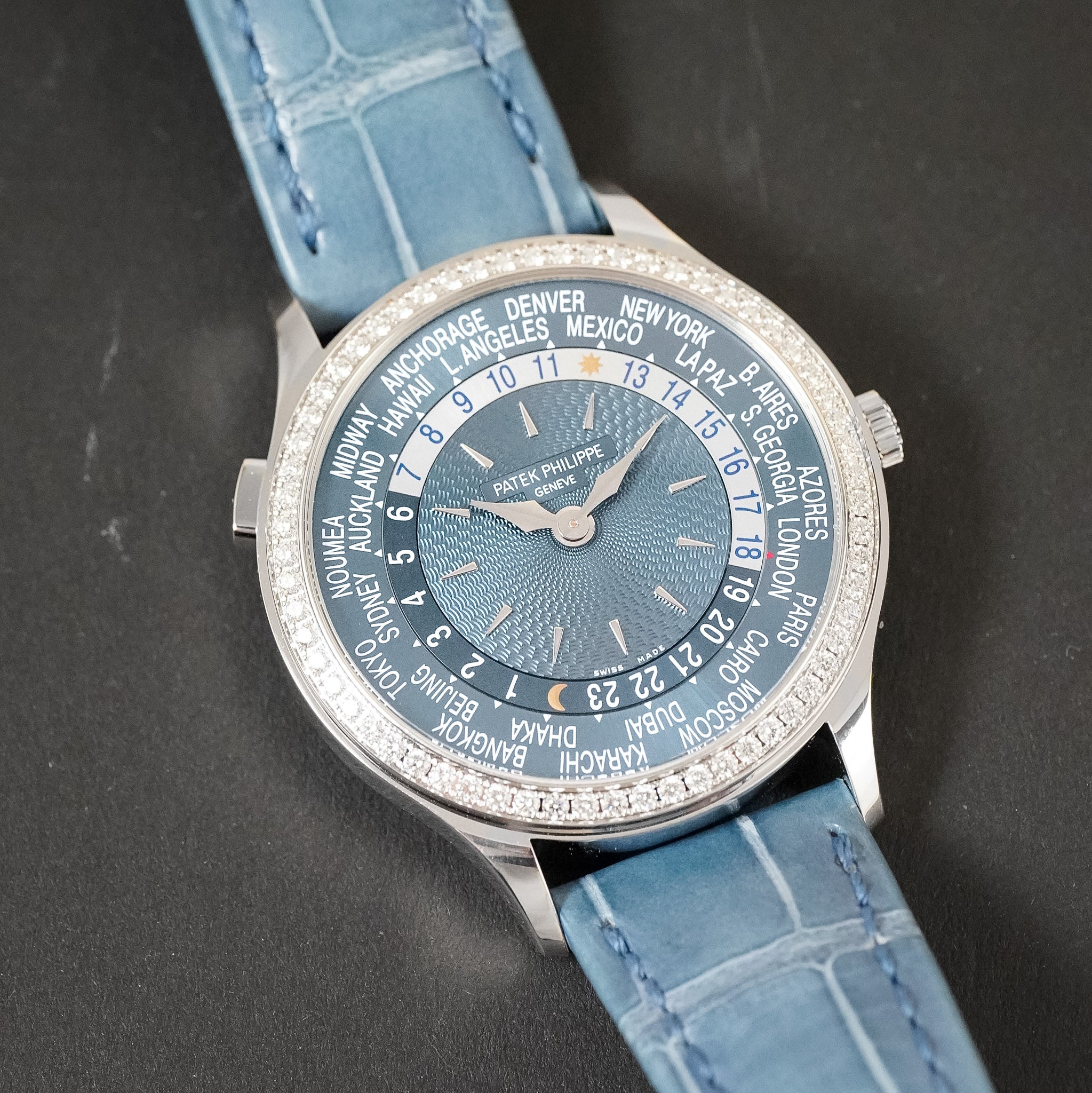 A light blue Patek Philippe World Time Watch with a ring of global cities around the dial. The watch is laying flat on a grey stone background. 