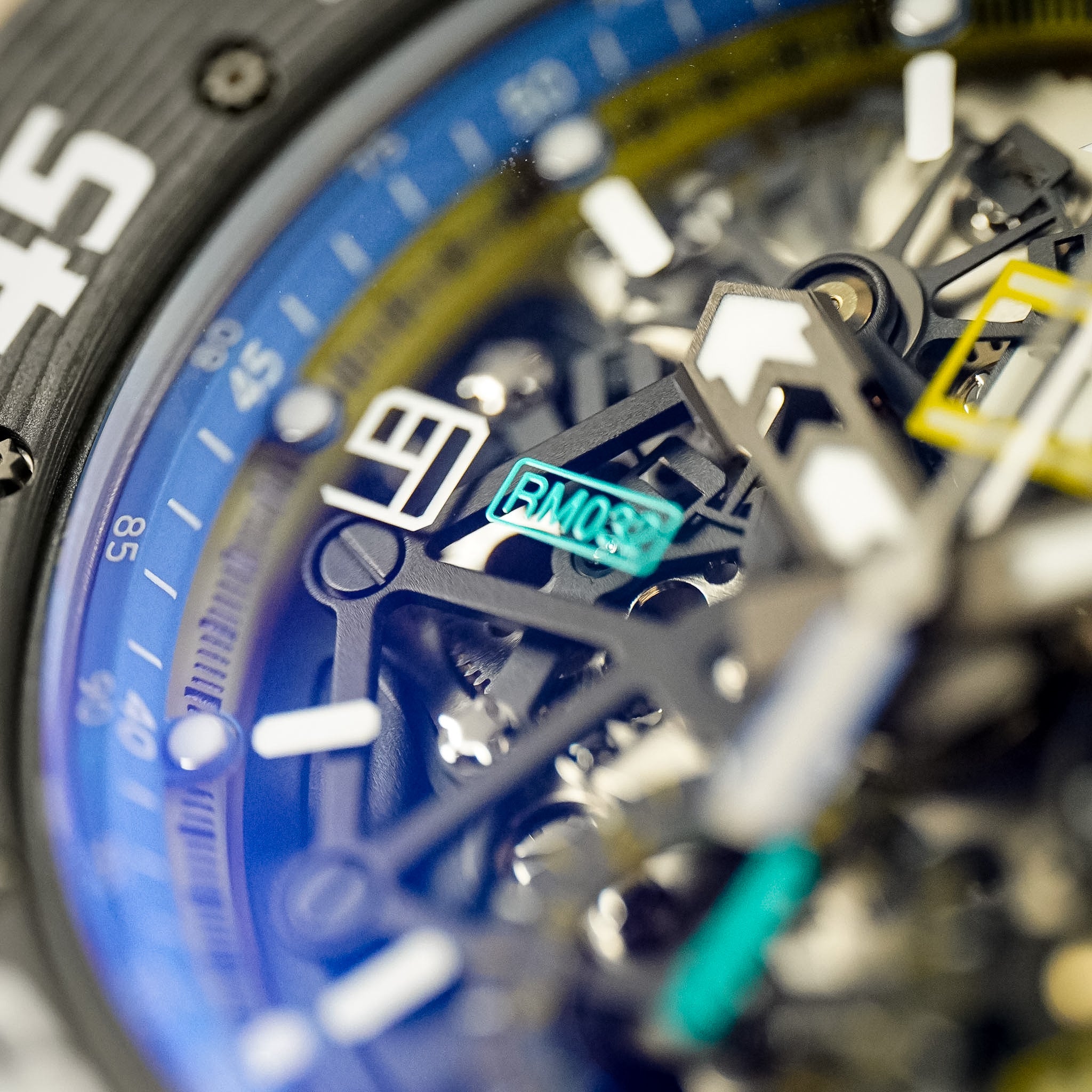 Close-up of a skeleton watch dial with a blue bezel, black and silver case elements, white hour markers, and a turquoise RM038 badge.