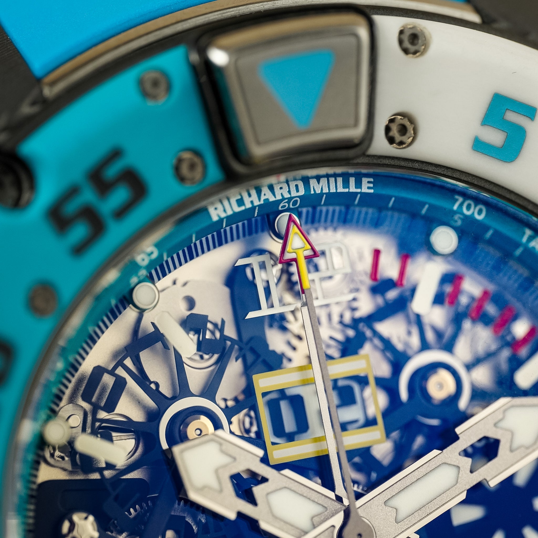 Close-up of a Richard Mille watch dial and bezel, showing a blue and white skeletonized face with silver hands and markers, plus turquoise and white sections on the outer ring.