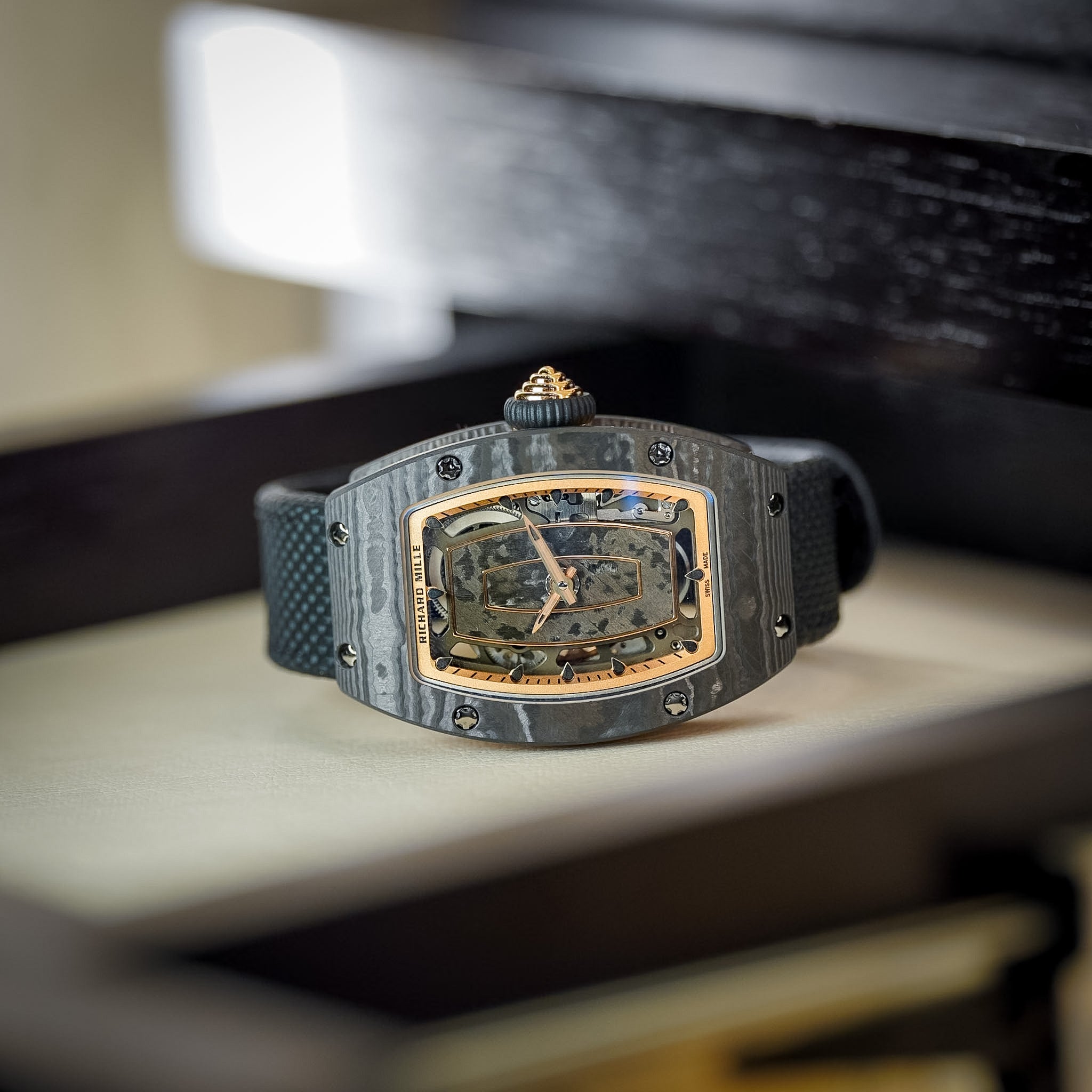 A luxury skeleton watch with a dark gray carbon fiber case, gold-tone case accents, and a black textured strap, resting on a light beige surface with a dark wooden background.