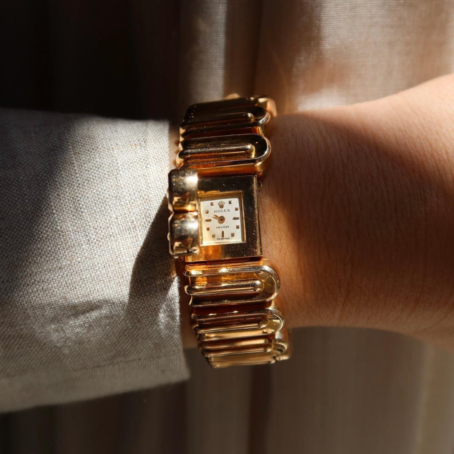 A gold-tone rectangular watch with a textured link bracelet, worn on a wrist. The watch has a white dial with gold-tone hour markers and hands.