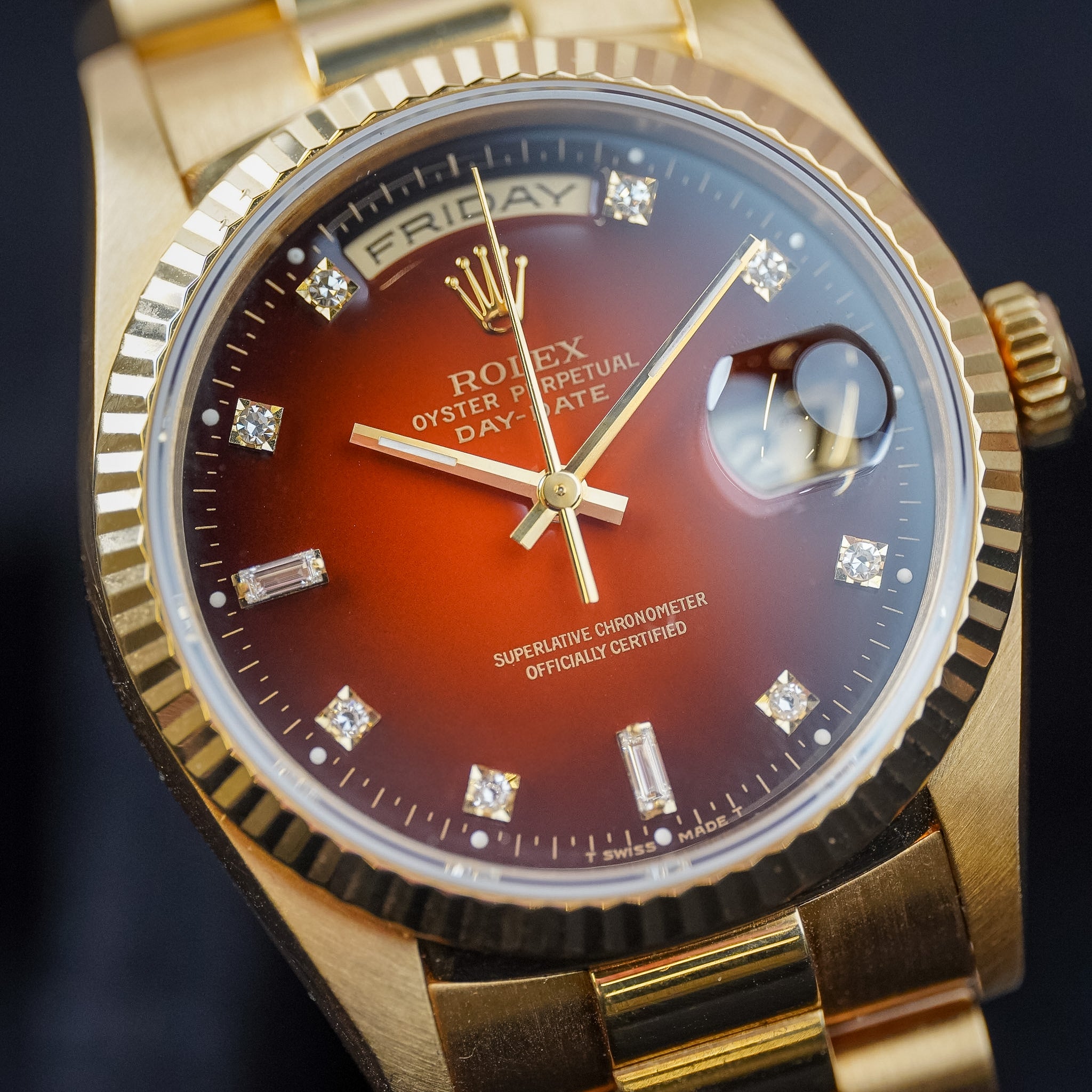Closeup of a yellow gold Rolex Day-Date watch with a burgundy ombé dial and diamond hour markers shot on dark surface
