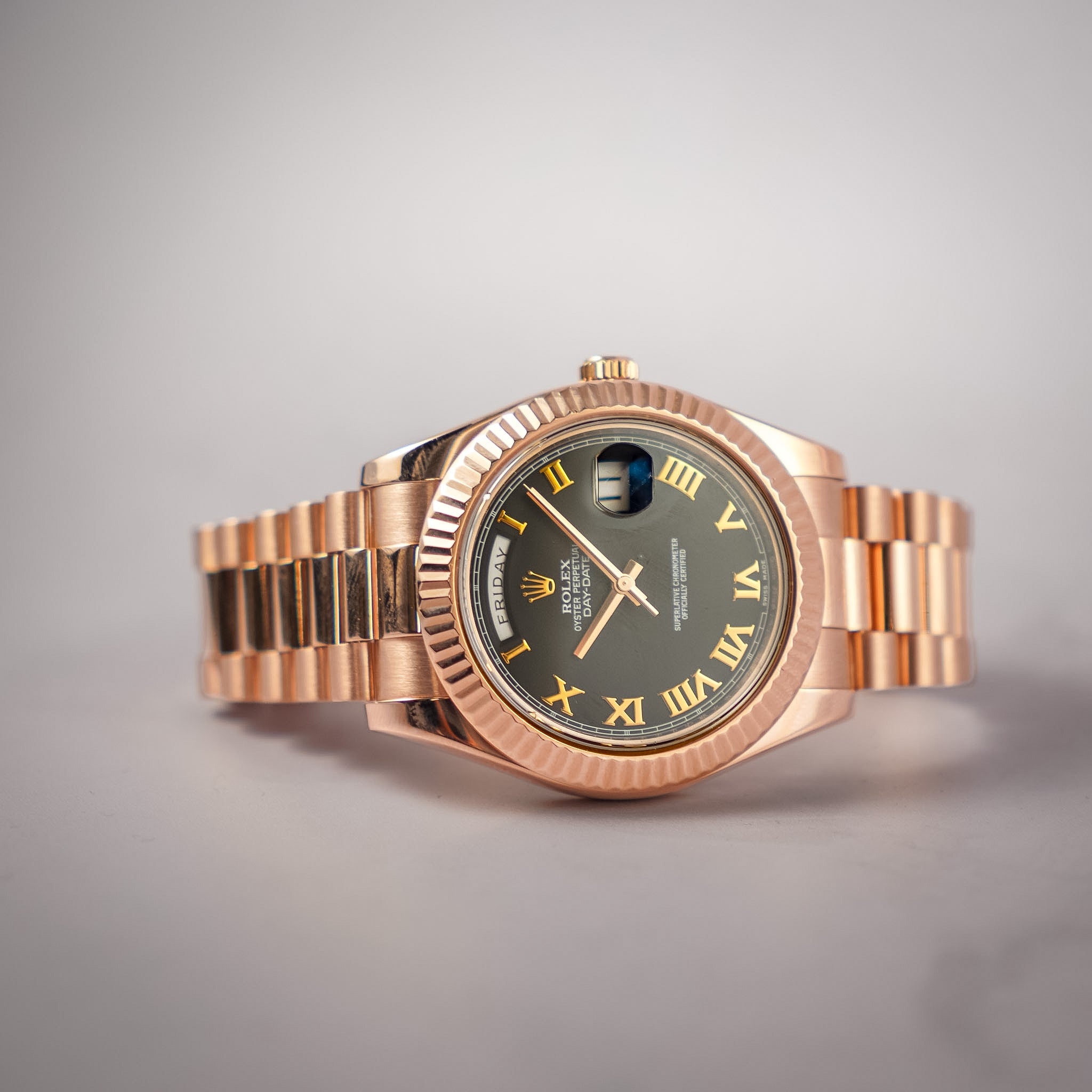 A rose gold Rolex watch with a black dial and Roman numeral hour markers laying on its side against a light gray backdrop.