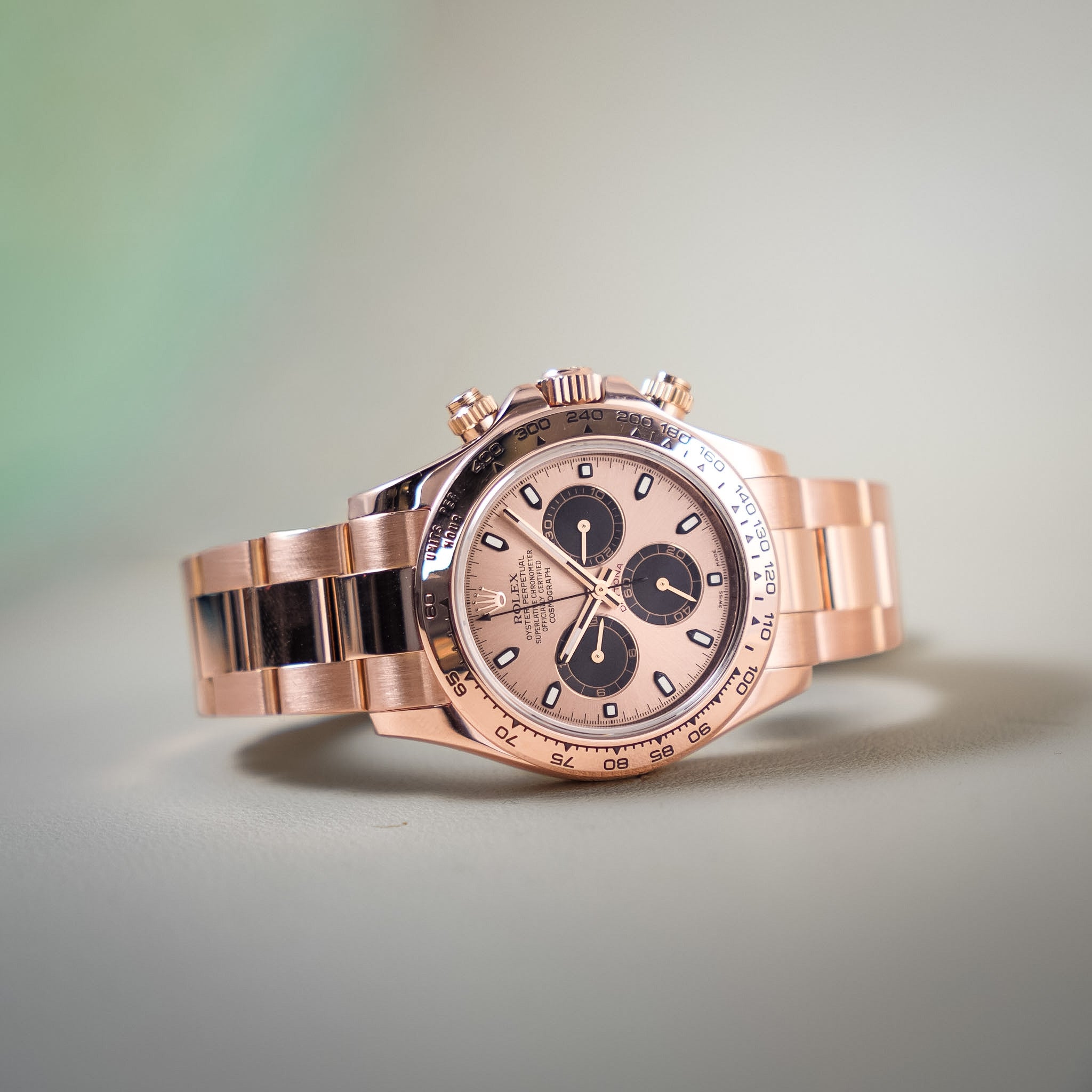 A luxury wristwatch with a rose gold-tone metal case and integrated bracelet, featuring a matching rose gold dial with black subdials and a tachymeter bezel, photographed on a soft light gray background.