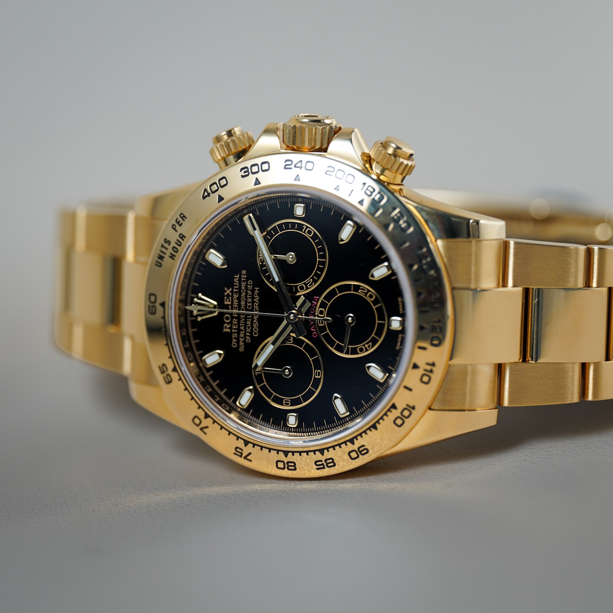 A gold-tone Rolex Daytona watch with a black dial and matching gold-tone Oyster bracelet, photographed close-up against a neutral gray background.