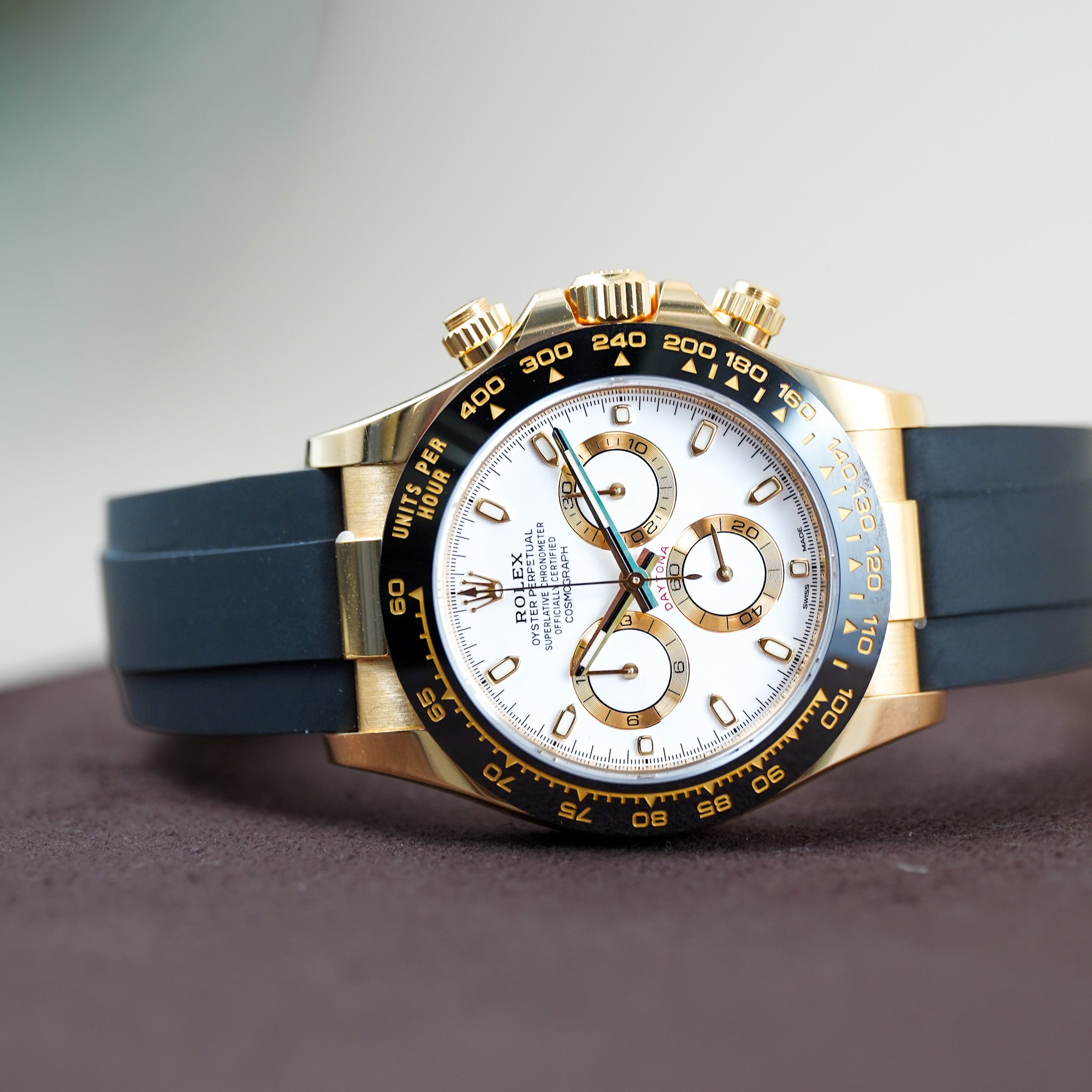 Rolex Daytona watch with a gold-tone case, white dial, blue rubber strap, and gold-tone bezel, displayed on a dark surface with a soft gray background.