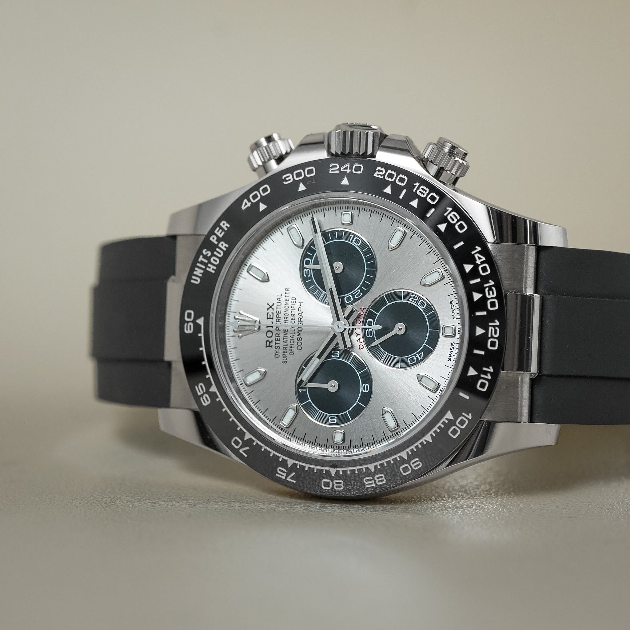 A Rolex Daytona watch with a silver-toned stainless steel case, a silver dial featuring blue subdials, and a black bezel and black rubber strap, shown on a neutral beige background.