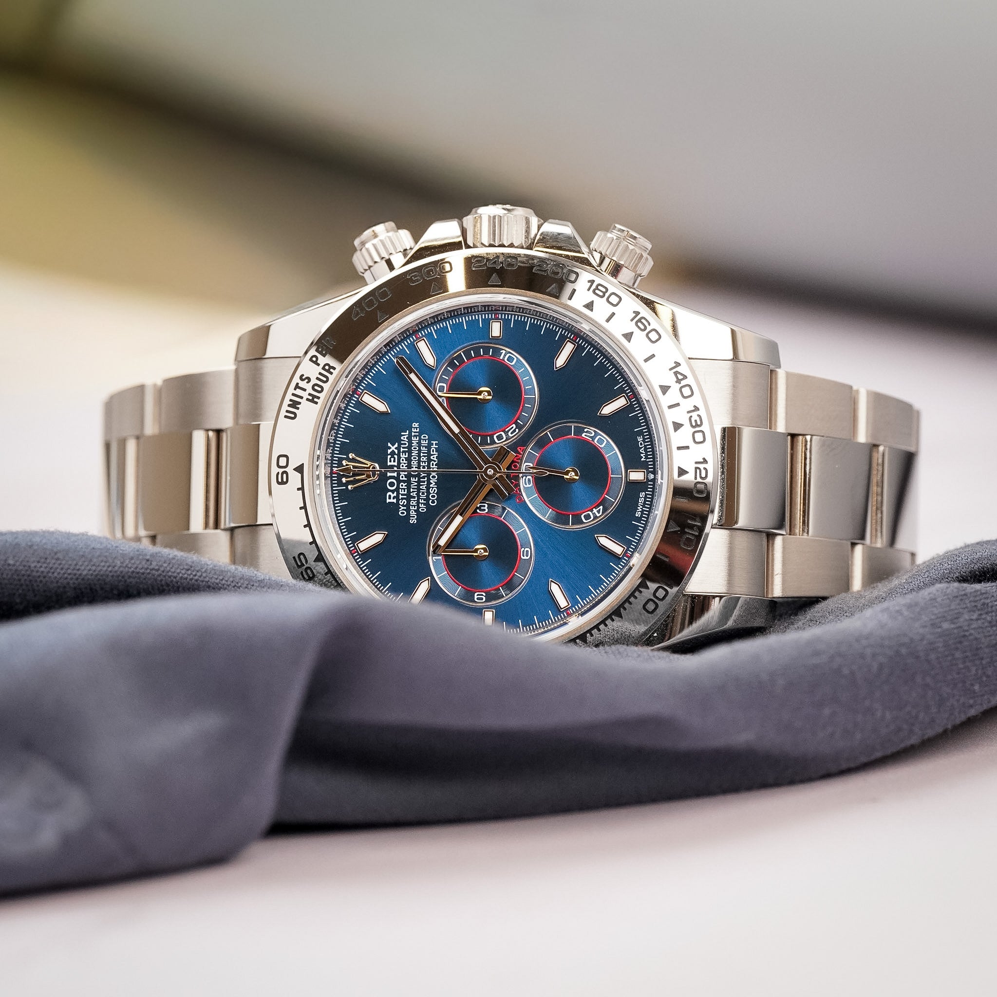 A white gold Rolex Daytona with a blue dial, red accents, and a white gold bracelet, shot laying on its side on a grey cloth and a white table
