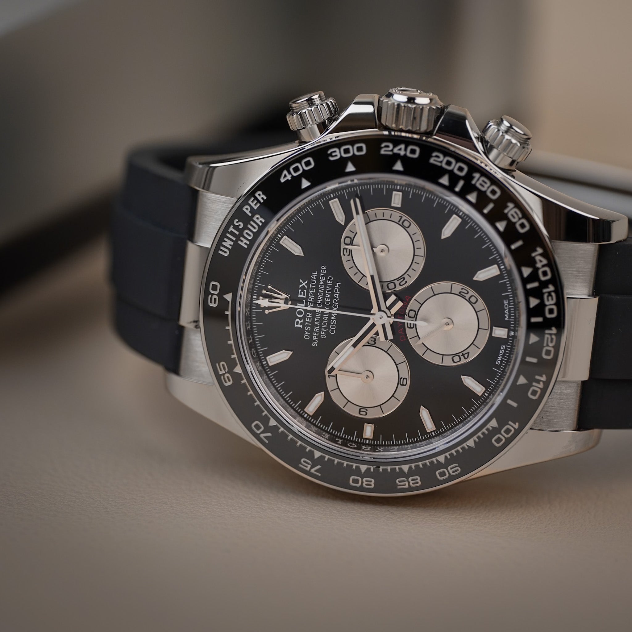 A Rolex Daytona in white gold with a black dial, silver subdials, a black ceramic bezel, and a black rubber strap. Shot lying sideways on neutral beige surface