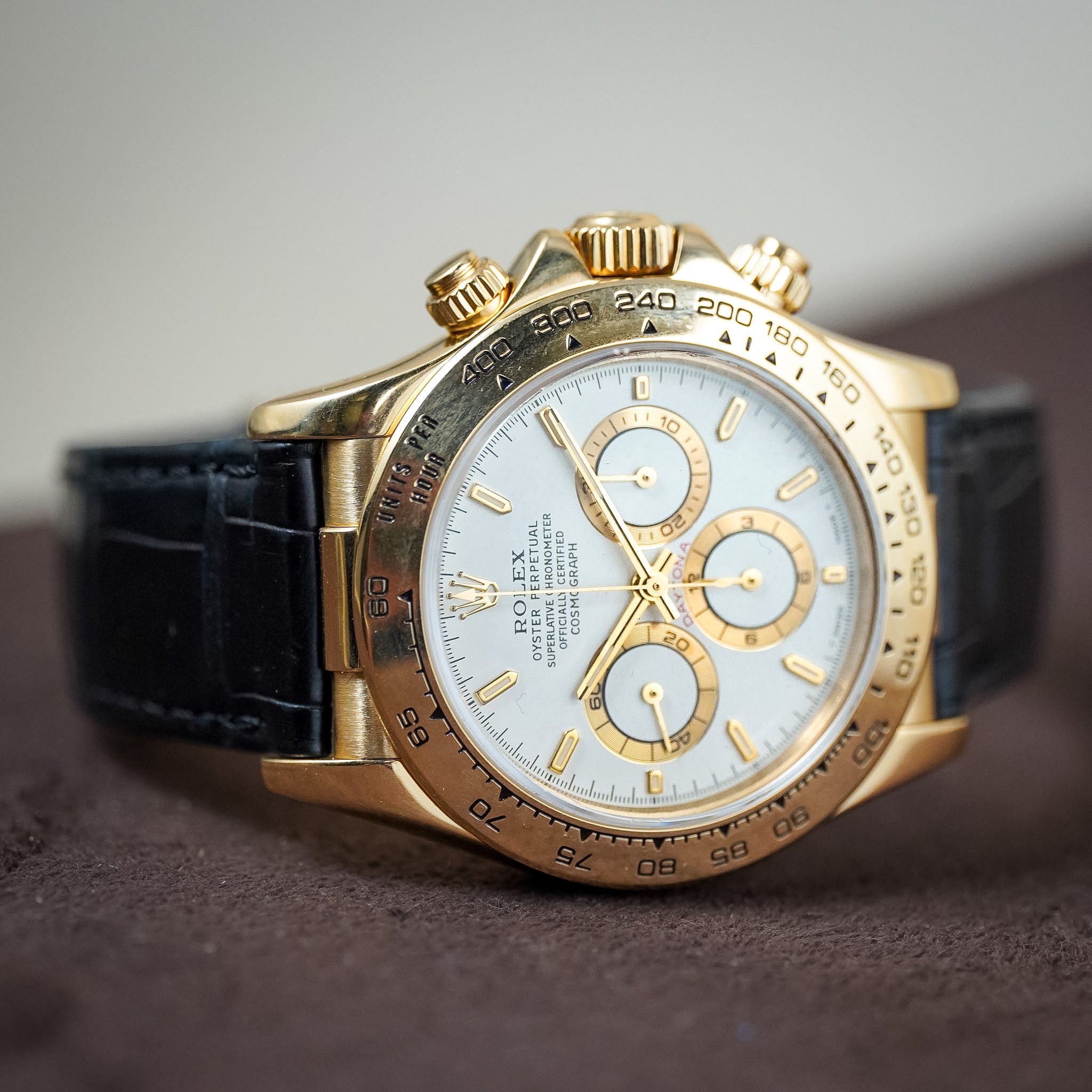 A Rolex Cosmograph Daytona wristwatch with a gold-tone case, white dial, and black leather strap, resting on a dark brown textured surface.