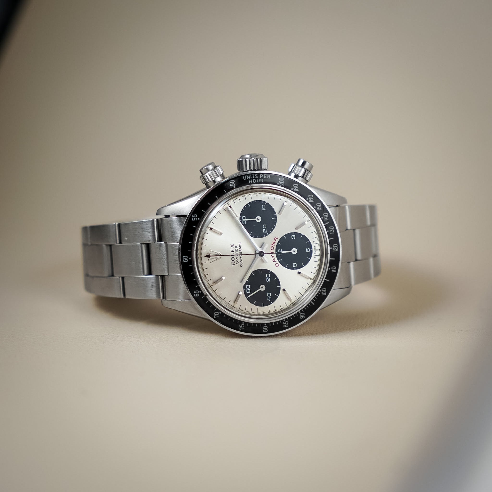 A stainless steel Daytona watch with a silver-tone metal bracelet, featuring a white dial with three dark blue subdials and a black tachymeter bezel, photographed on a neutral beige background.