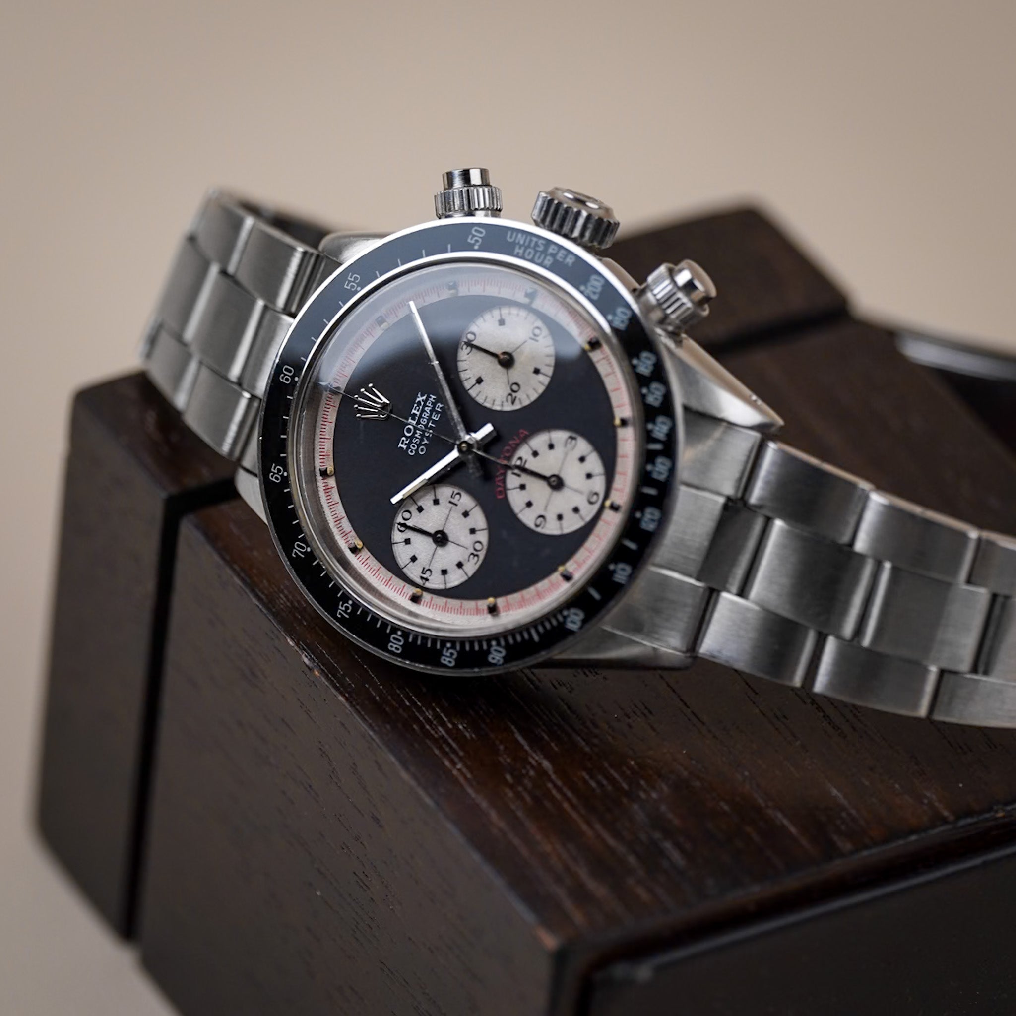 A black dial vintage Rolex Daytona in steel with a black bezel, white subdials, and red accents, lying sideways on a dark wooden box and neutral beige background