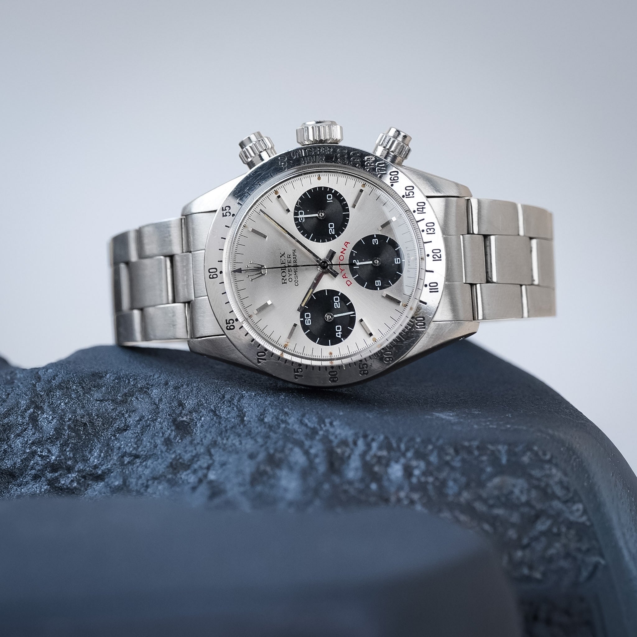 A vintage Rolex chronograph watch with a steel case, steel bezel, silver dial with black subdials, and a steel bracelet, shot resting sideways on a textured blue form with a light grey background.