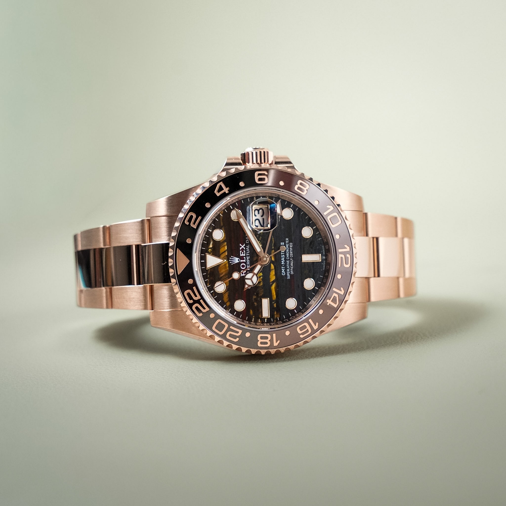 A Rolex GMT-Master II watch with a rose gold-tone metal bracelet and case, featuring a black dial and a two-tone brown and rose gold bezel, set against a soft light green background.