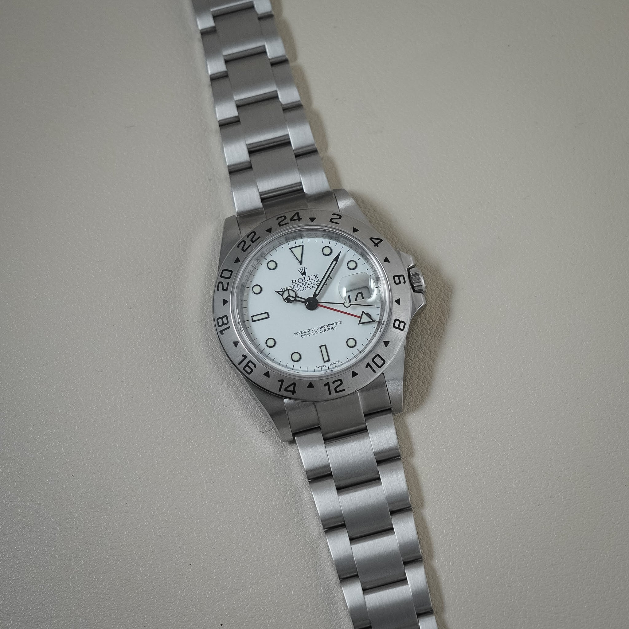A Rolex Explorer II watch with a silver stainless steel Oyster bracelet, a white dial, and a black 24-hour bezel, photographed on a neutral light beige background.