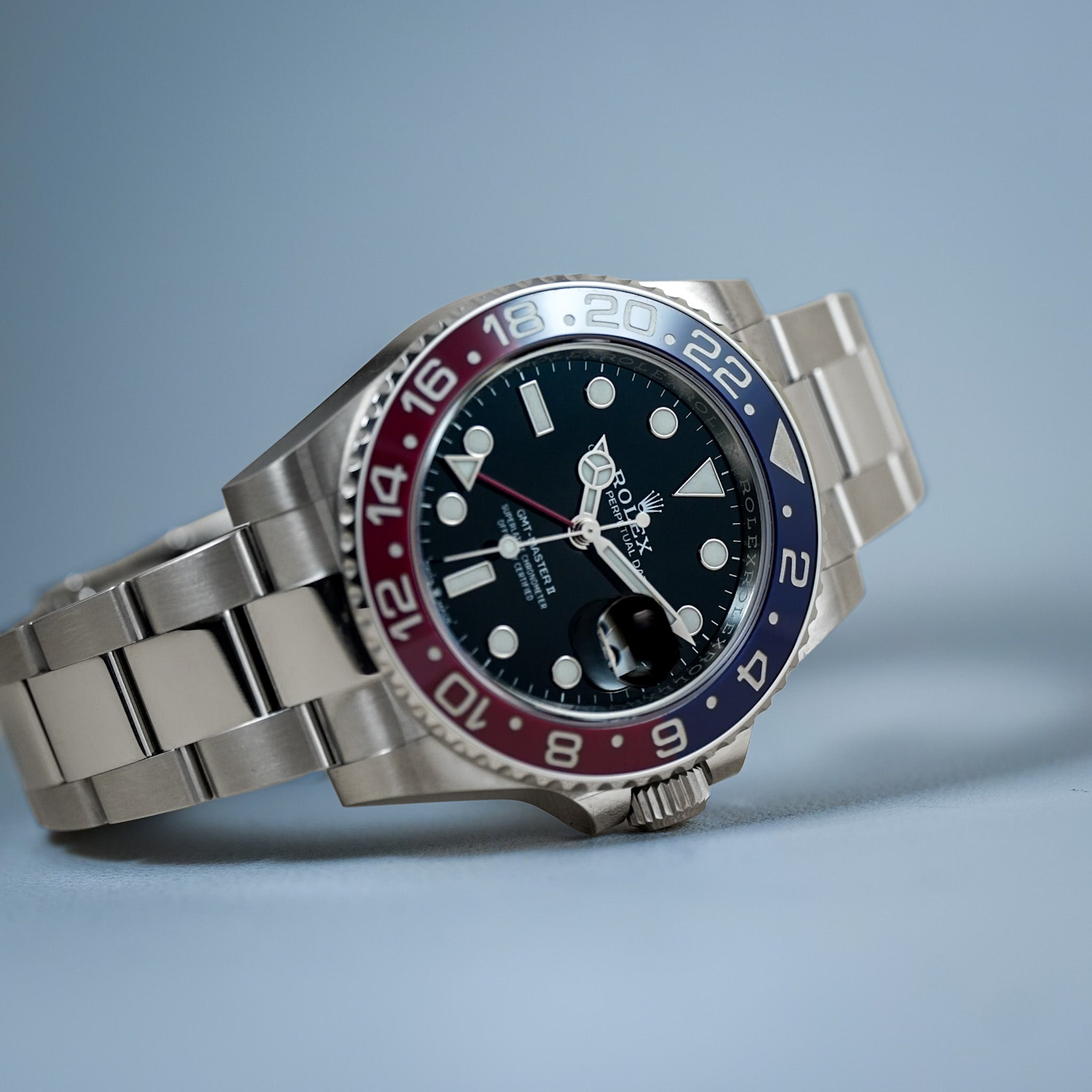 A stainless steel Rolex GMT-Master II with a black dial and a red and blue bezel laying sideways on a light blue background.