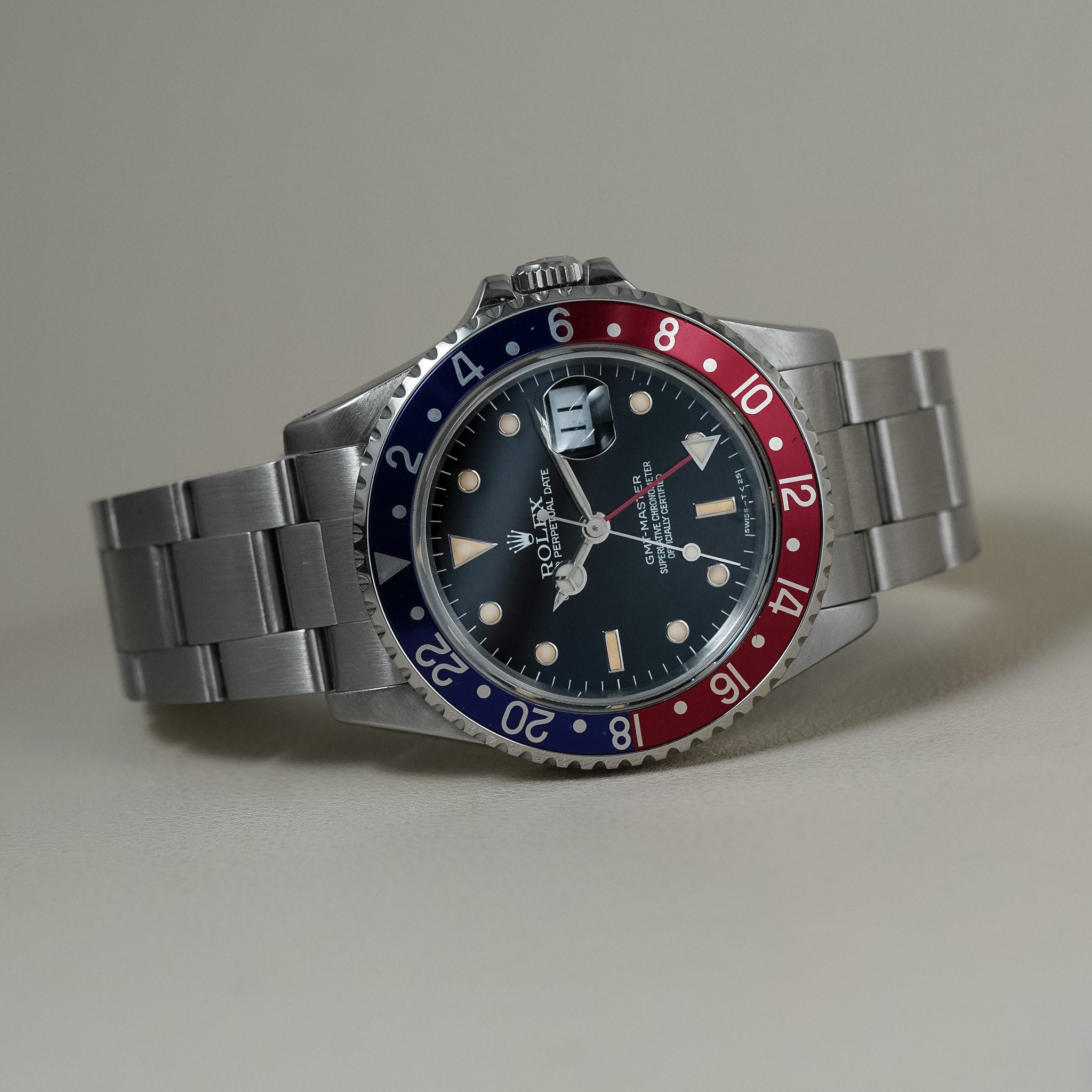 A Rolex GMT-Master watch with a stainless steel bracelet and a distinctive red and blue bezel, displayed on a neutral beige background.