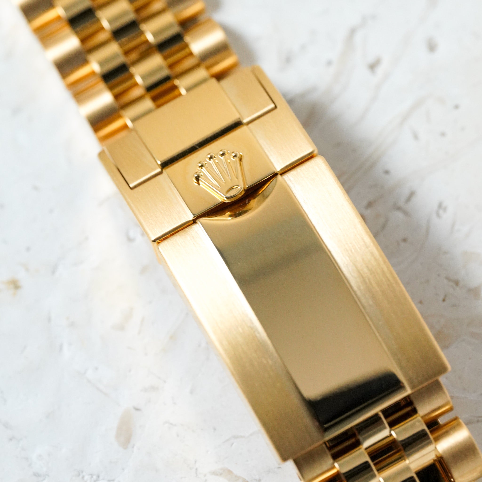 Close-up of a gold-tone metal bracelet watch band clasp with a crown logo, shown on a light textured surface.