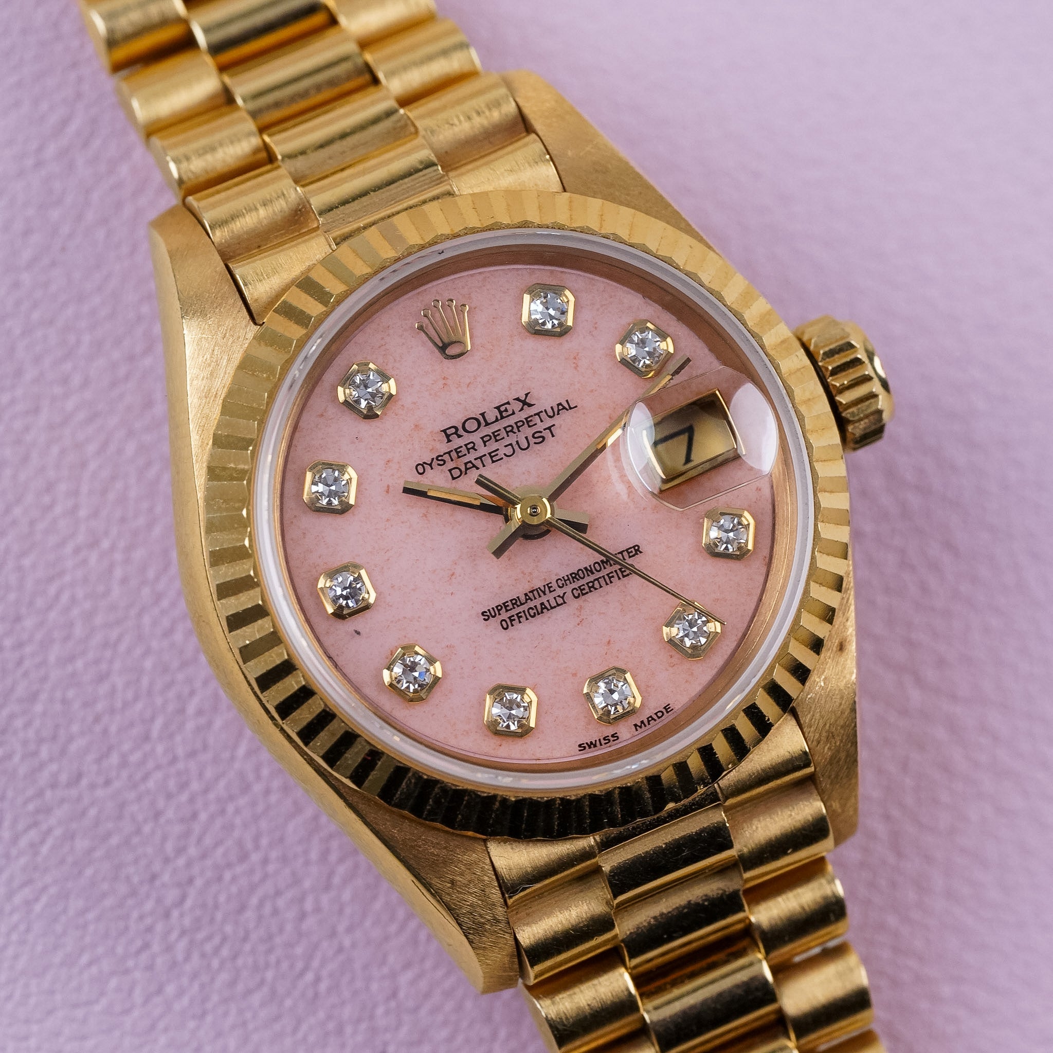 A yellow gold Rolex Lady Datejust with a fluted bezel, pink coral dial, diamond hour markers, gold hands, and a date window at 3 o'clock. Shot resting flat against a light pink surface.