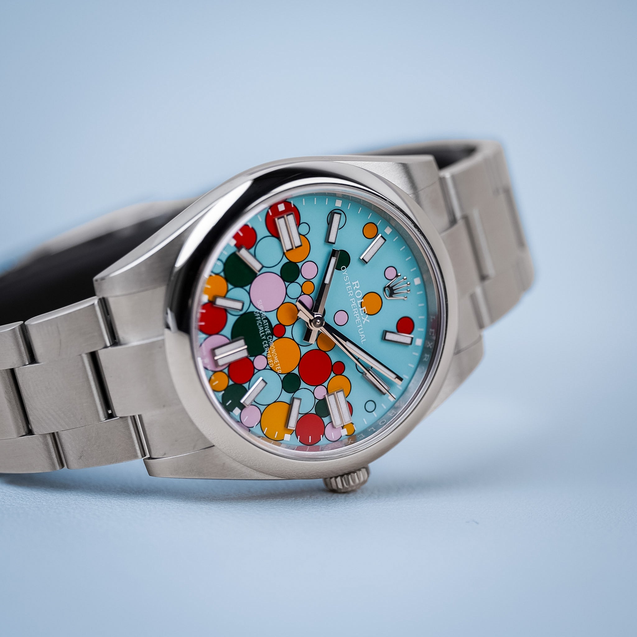 A stainless steel Rolex watch with a colorful blue dial featuring accents of red, yellow, pink, and green. Shot resting sideways on a solid light blue background.