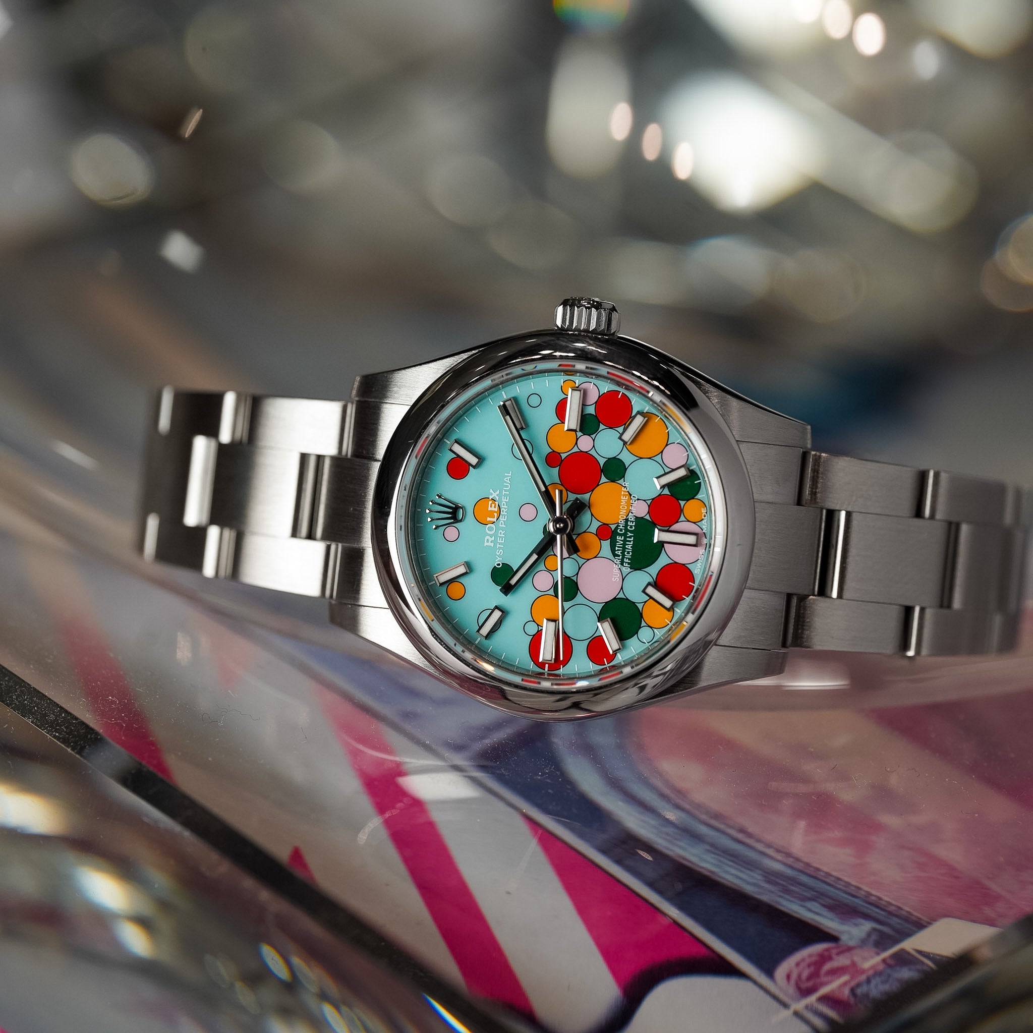 A stainless steel wristwatch with a colorful dial featuring a playful pattern of red, yellow, green, and orange shapes on a light blue background, resting on a reflective surface.