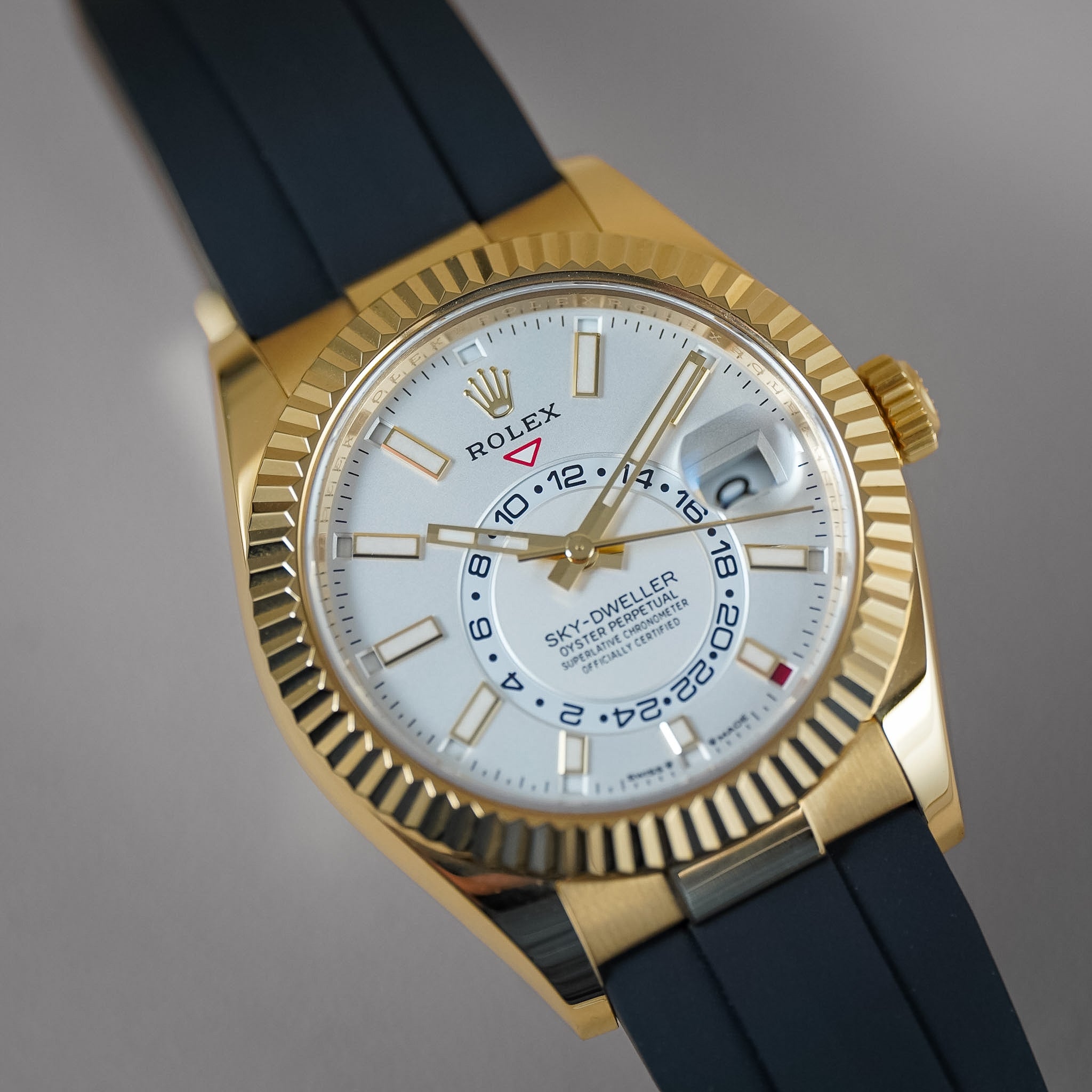 A yellow gold Rolex Sky-Dweller wristwatch on a black rubber strap. Shot on a neutral grey background.